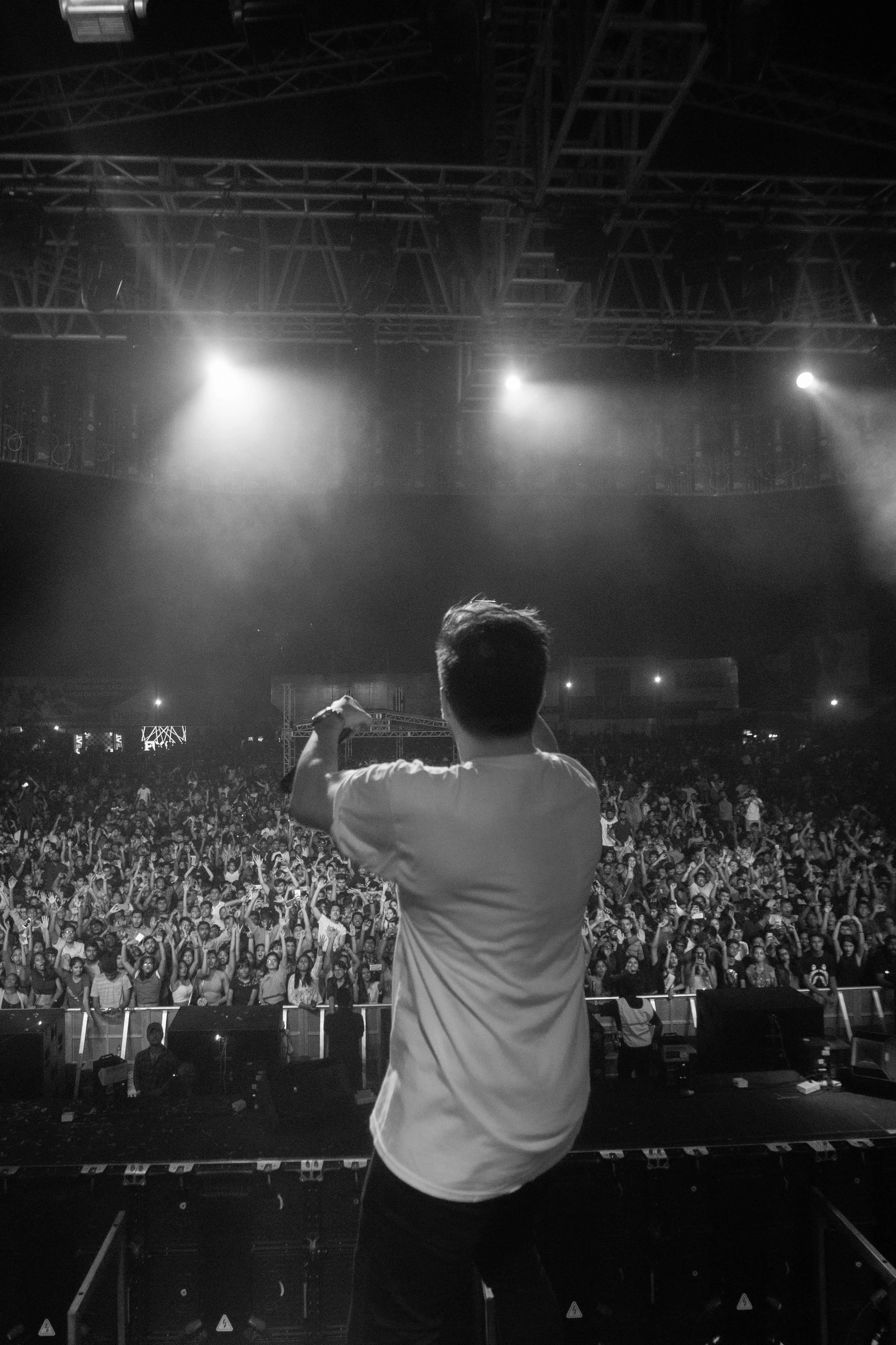 A DJ or performer on stage in front of a large crowd at a concert or club, with stage lighting overhead.