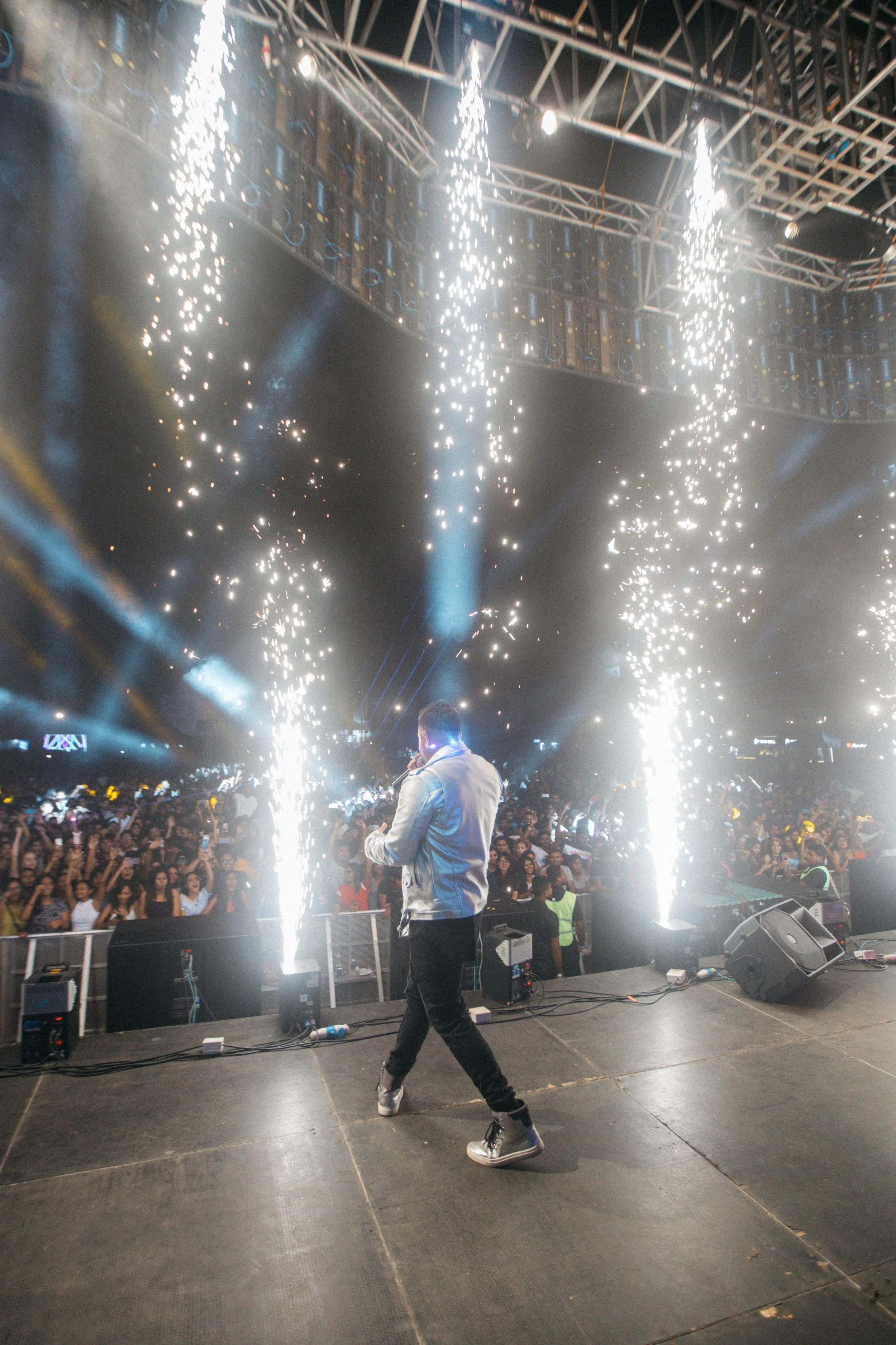 A performer on stage with pyrotechnics and bright lights, facing a large crowd at a concert or music event.