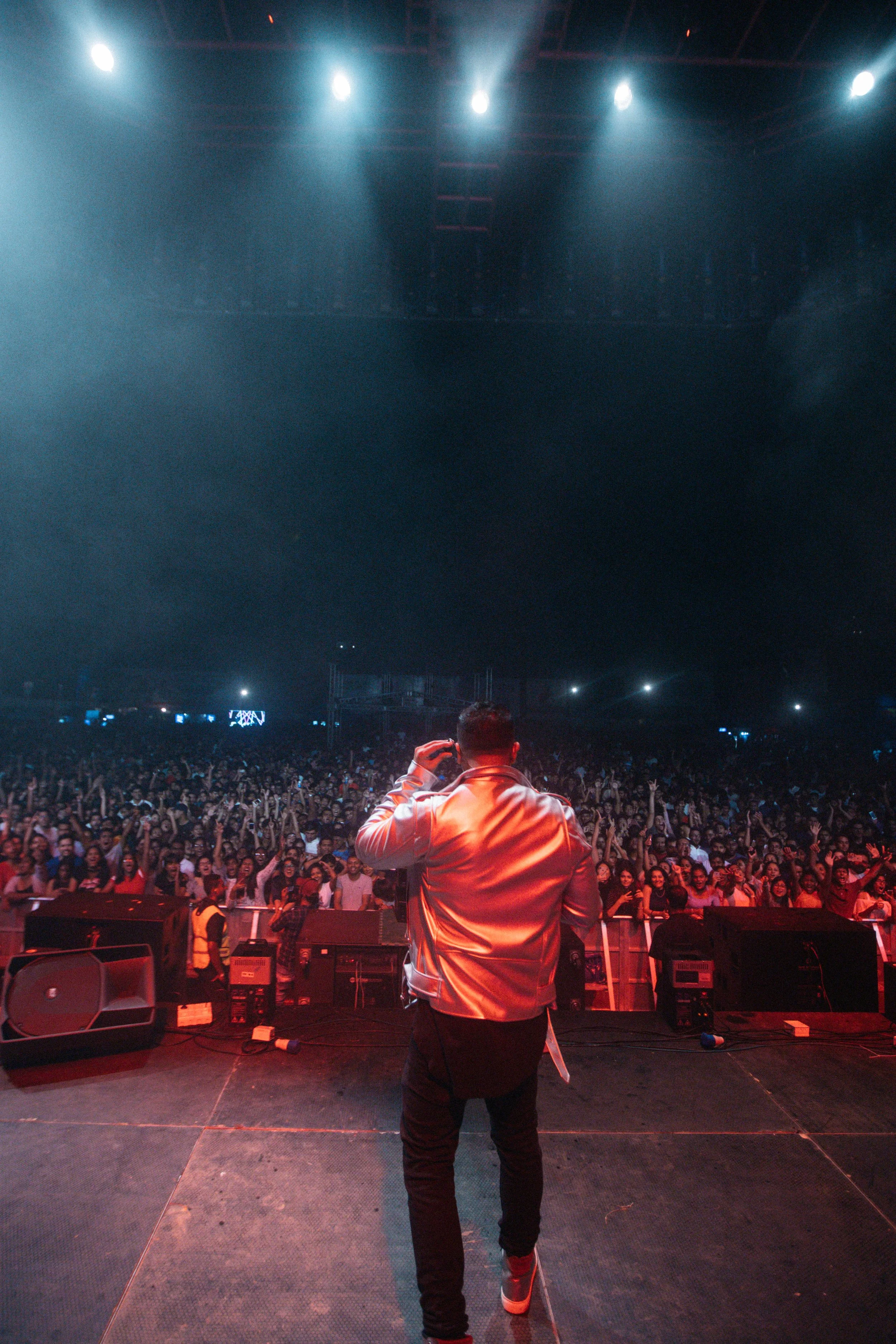 Performer on stage in front of a large audience at a concert venue with bright stage lights.