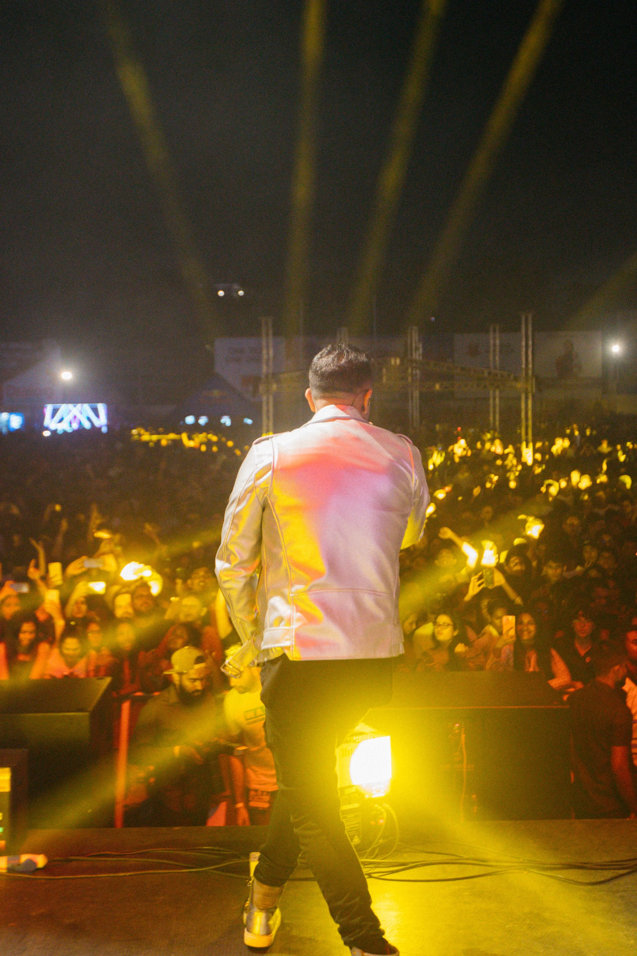 Performer on stage with his back to the camera, facing a large audience at a concert or event, illuminated by yellow stage lights.