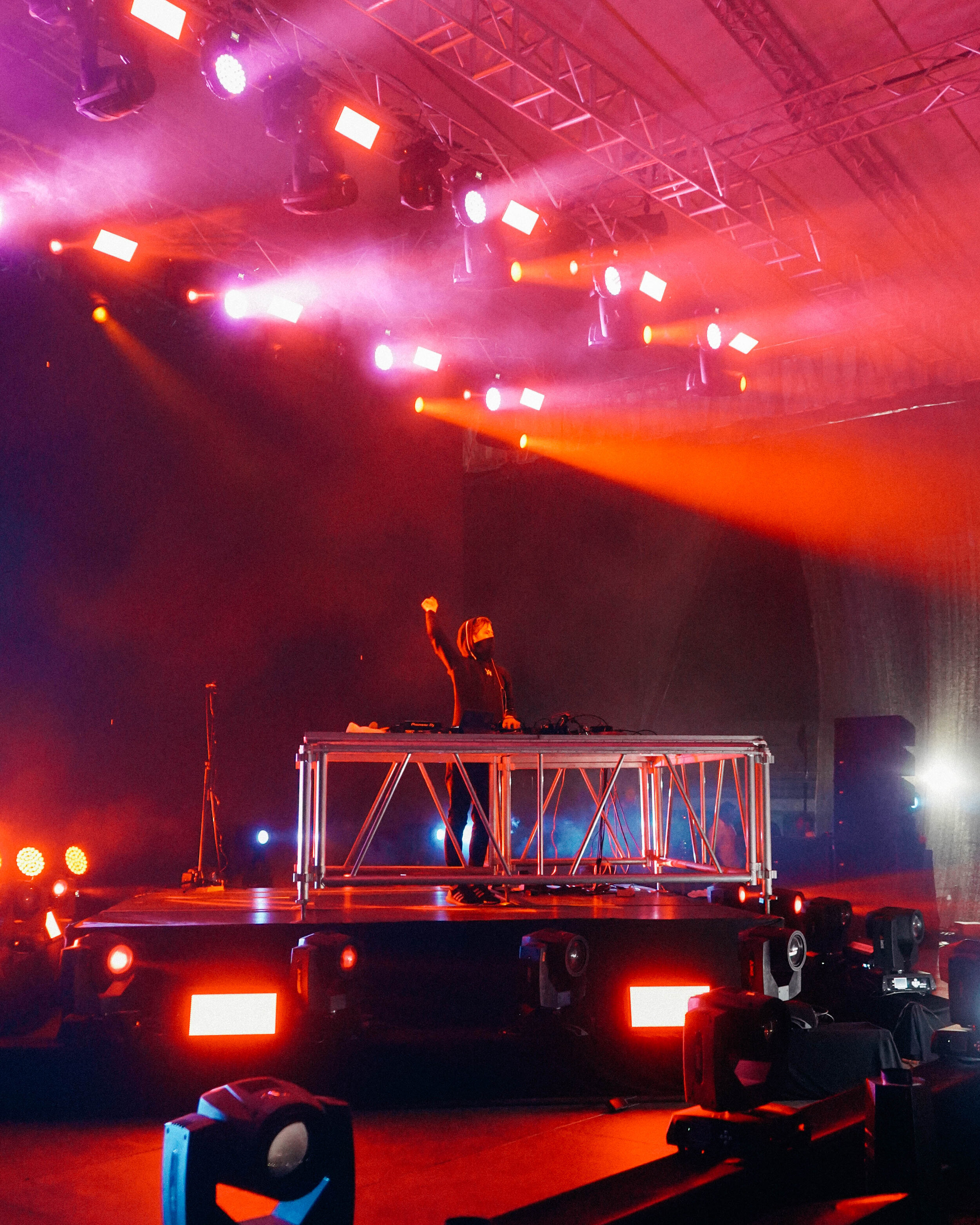 A DJ wearing a face mask and hoodie standing behind a DJ console on stage with colorful lights and fog, raising their right fist during a concert or music event.