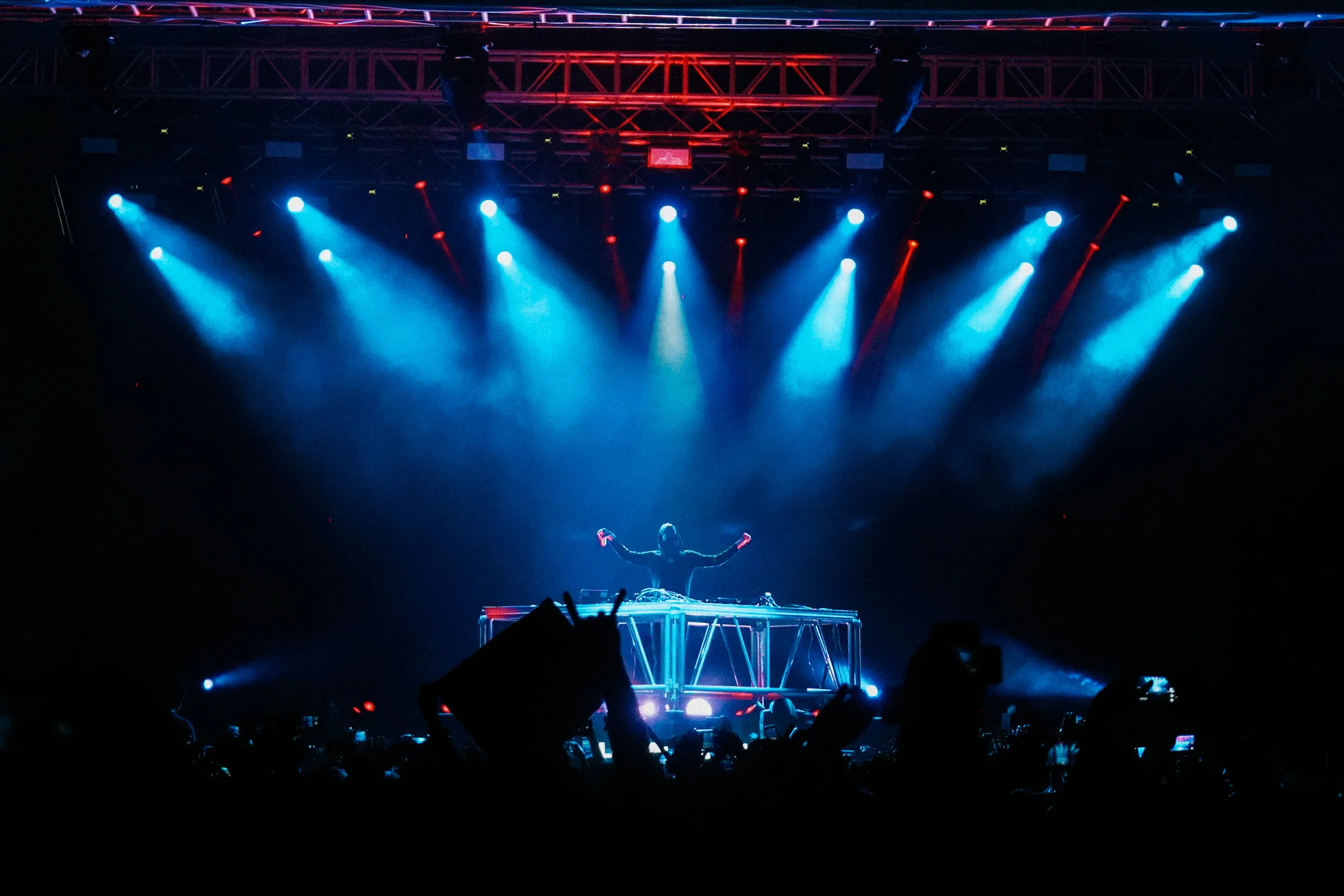 A DJ performing on stage with blue and red stage lights shining down, and a crowd cheering in the foreground.
