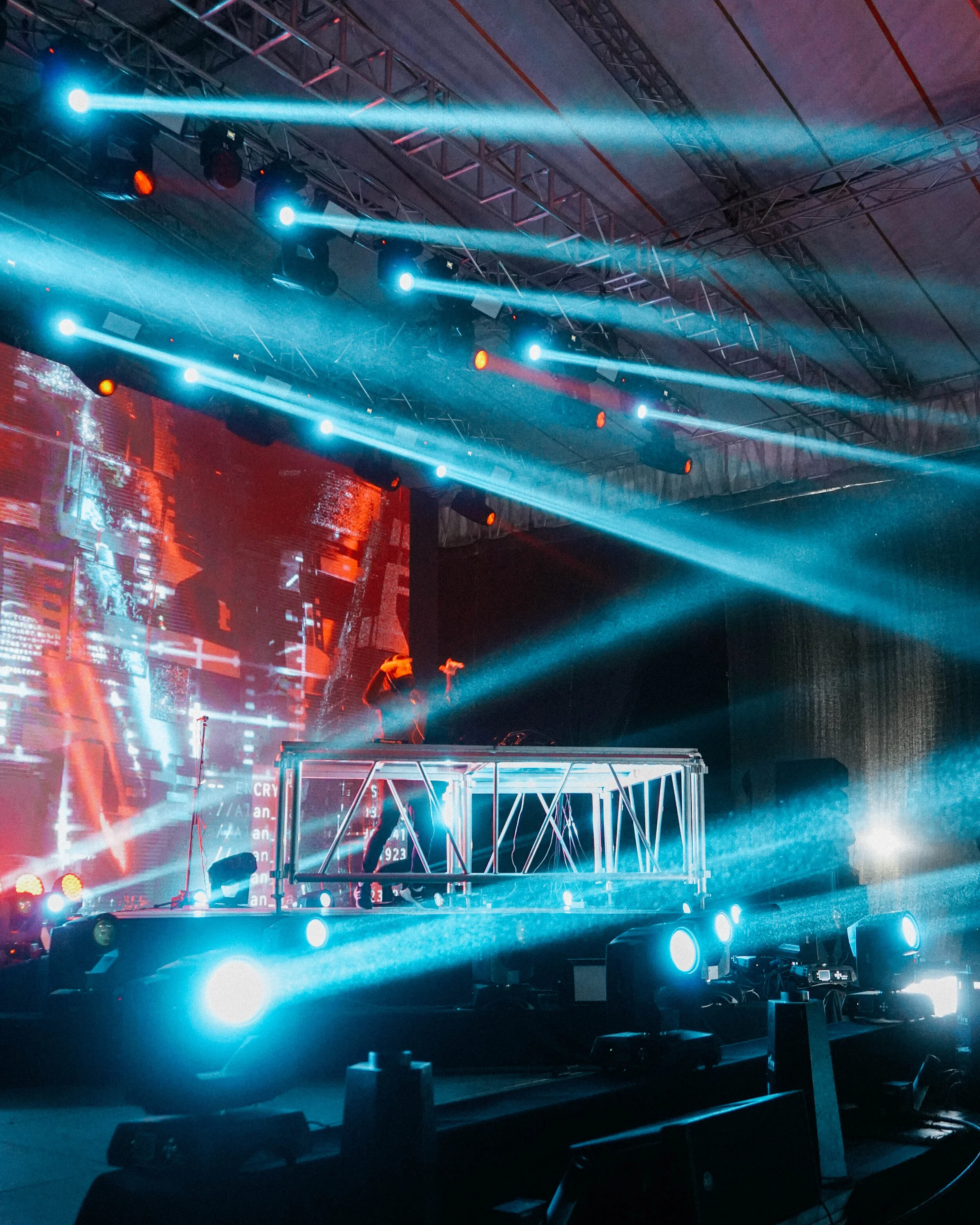 A DJ performing on a stage with colorful light beams and a digital screen background at a concert or music event.