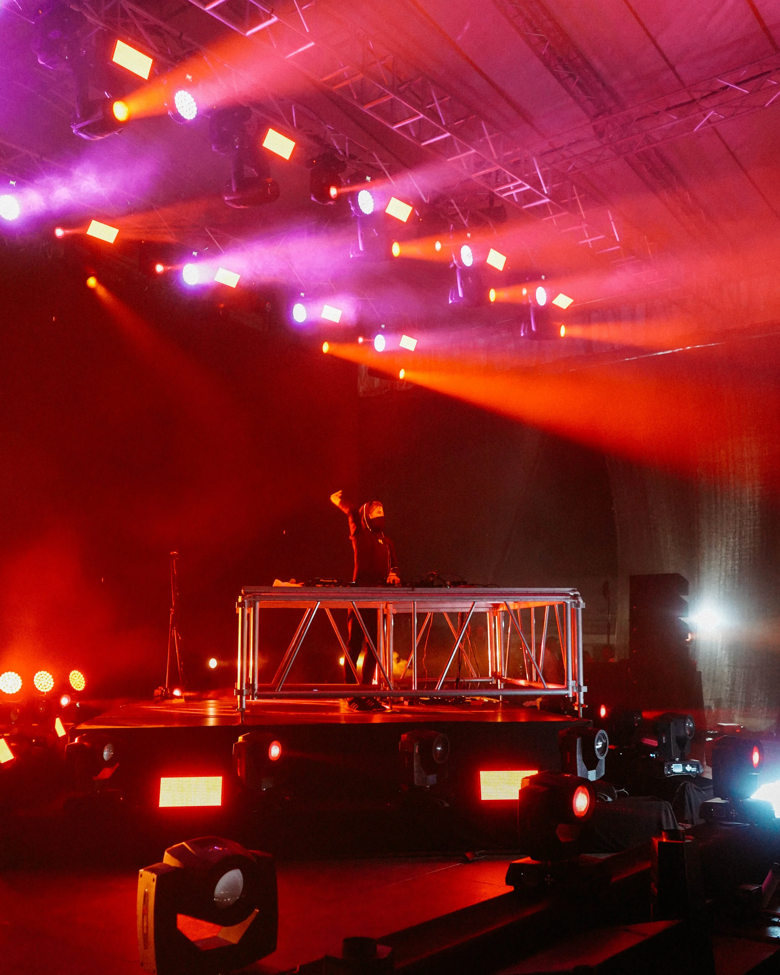 A DJ is performing on a stage illuminated with red and purple lights, standing behind DJ equipment on a raised metal platform, raising one arm in the air.