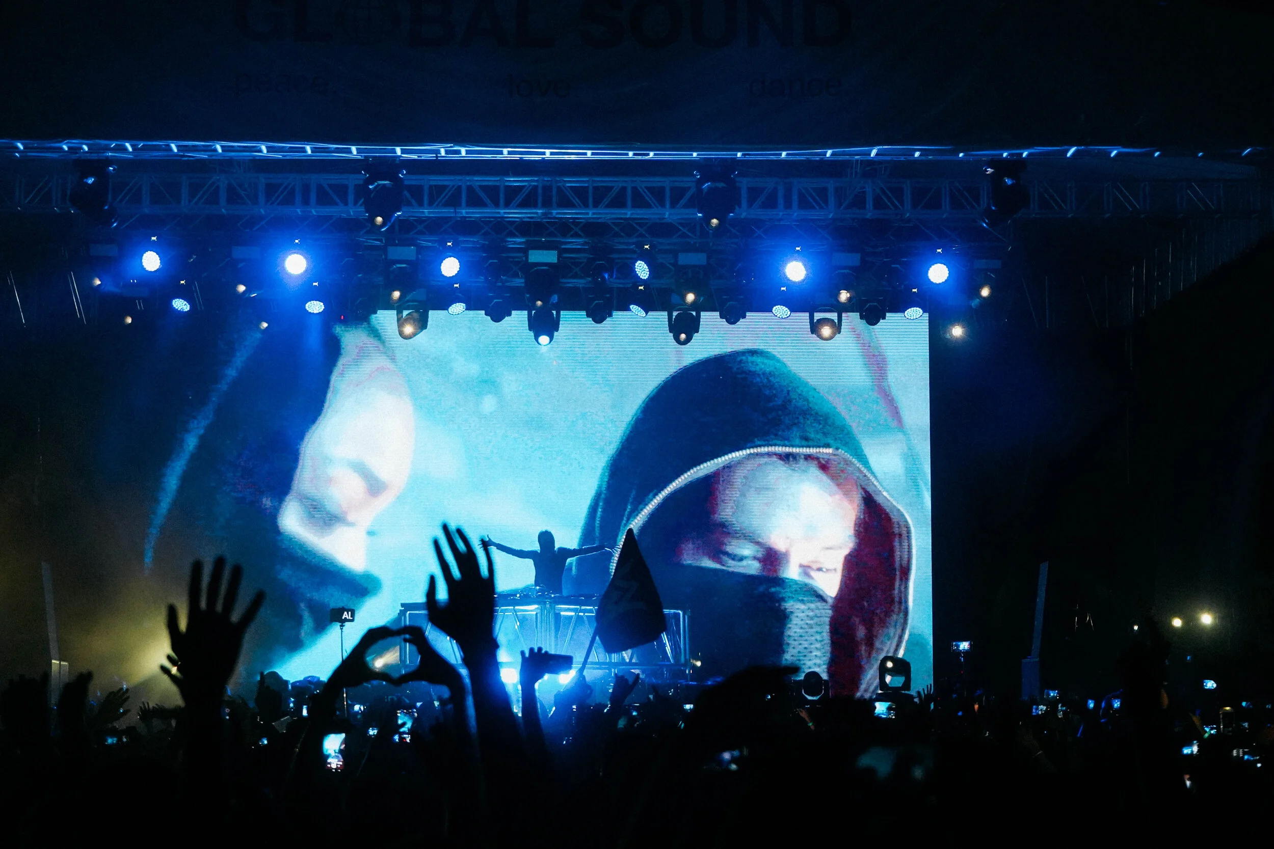 Concert scene with a large screen displaying a person wearing a hoodie and face mask, stage lights overhead, and a crowd with hands raised.