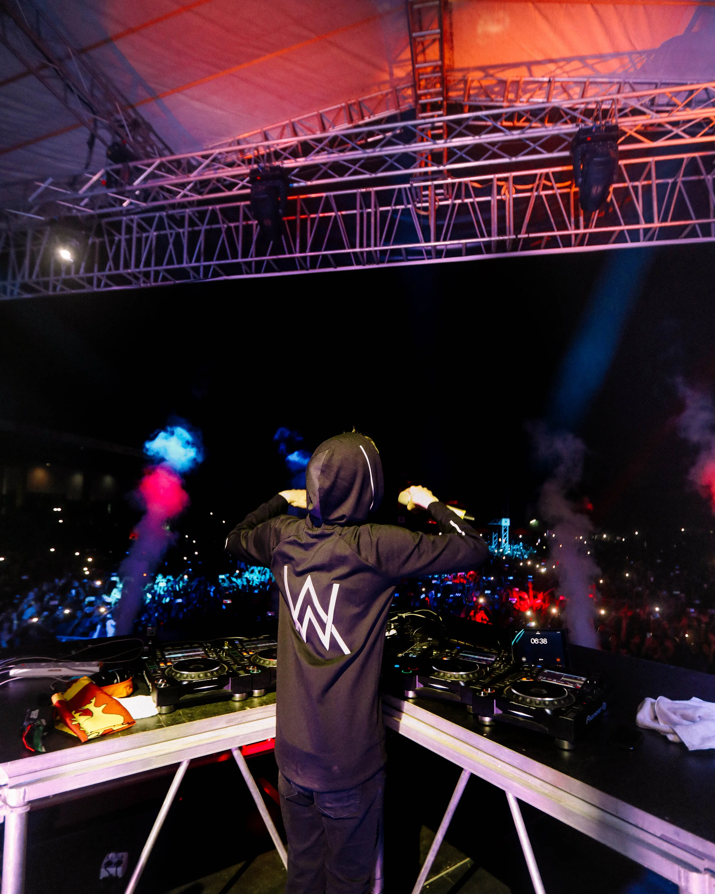 DJ performing on stage at a concert with a large crowd in the background, colorful smoke effects, and bright lighting overhead.