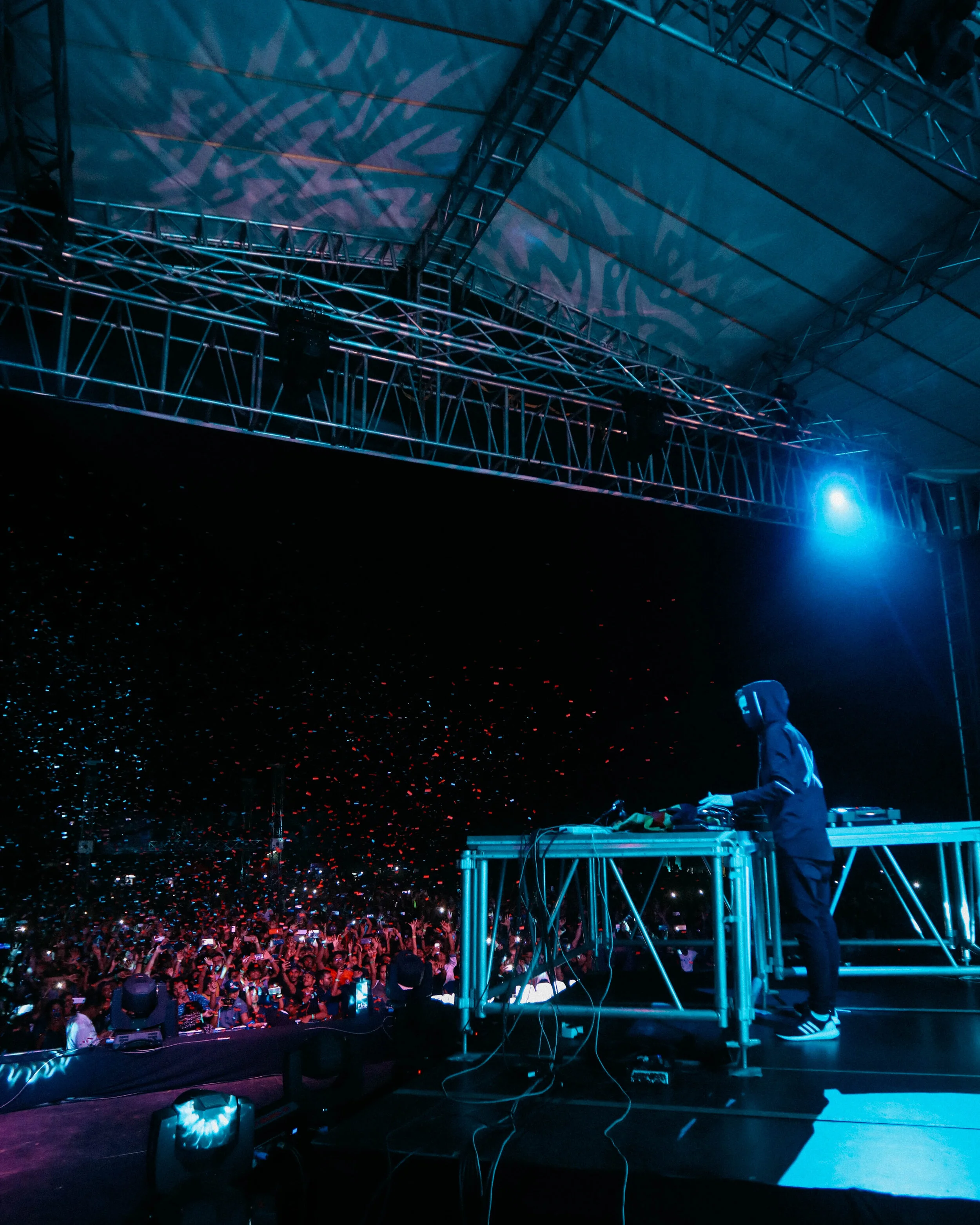 A DJ wearing a hoodie and headphones performing on a stage with a large crowd at night, illuminated by blue and purple stage lights.