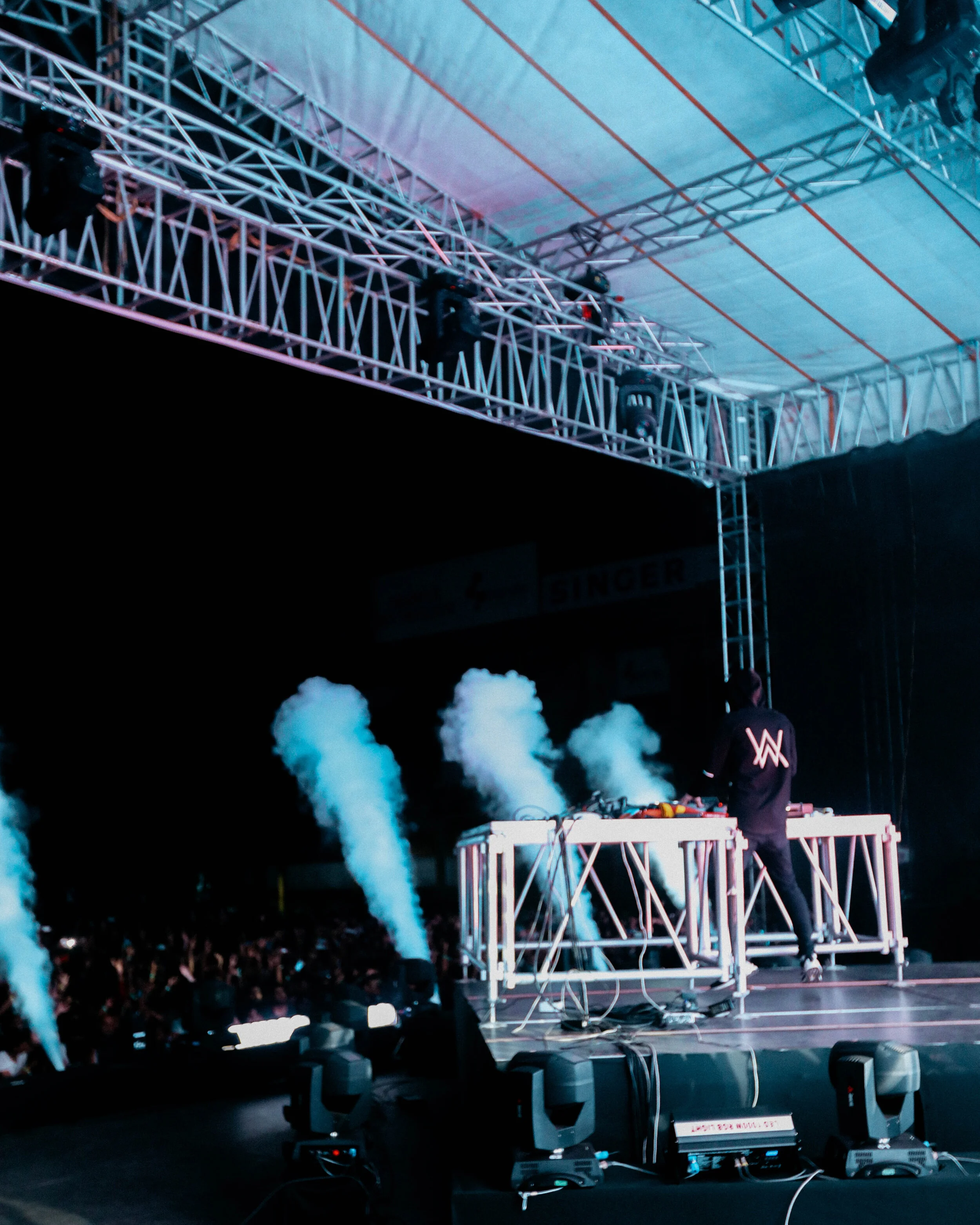 A DJ performing on stage at a concert with smoke effects and crowd in the background, with stage lighting and equipment.