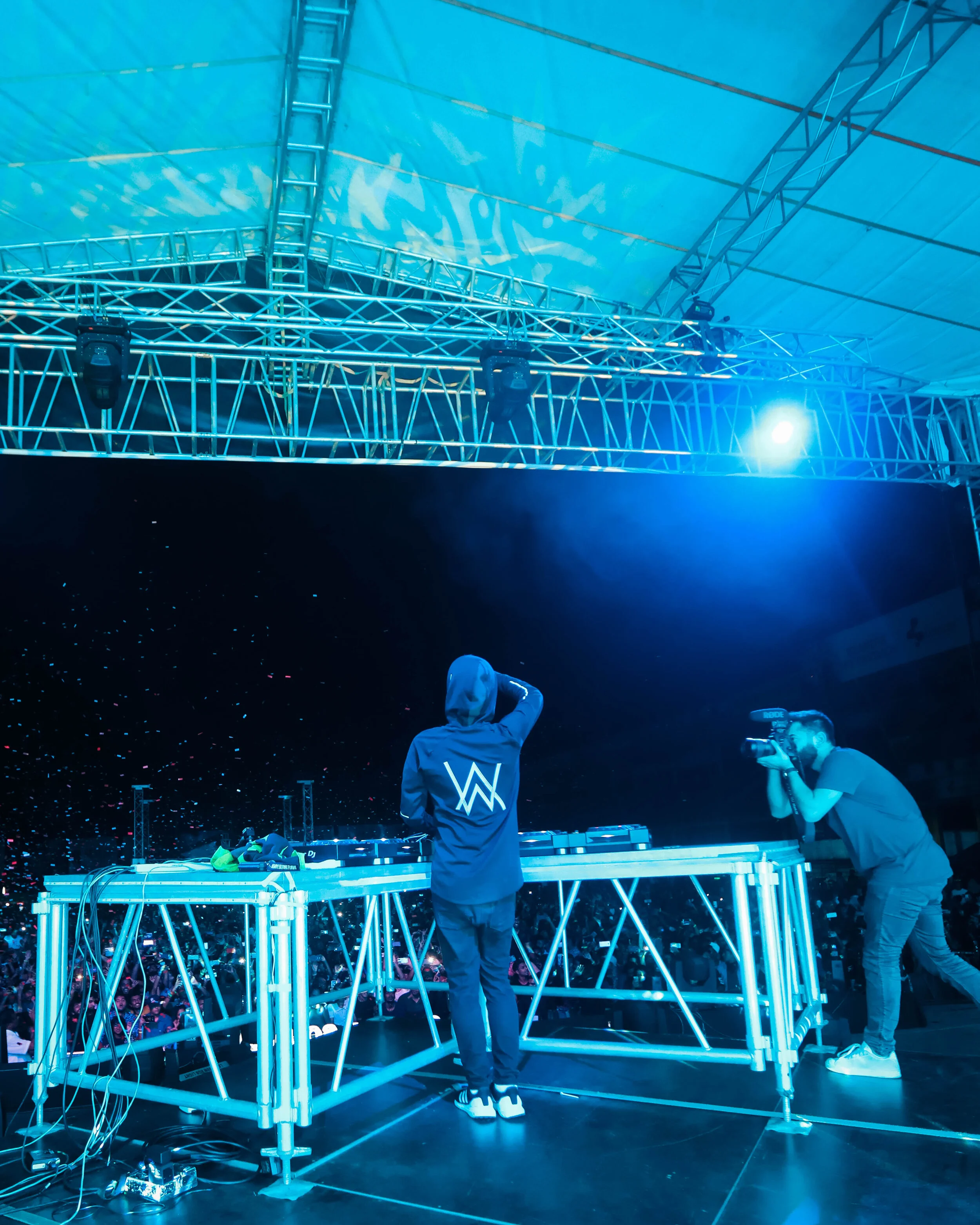 DJ performing on stage at a concert with a photographer capturing the moment, illuminated by blue stage lights, with a crowd in the background.