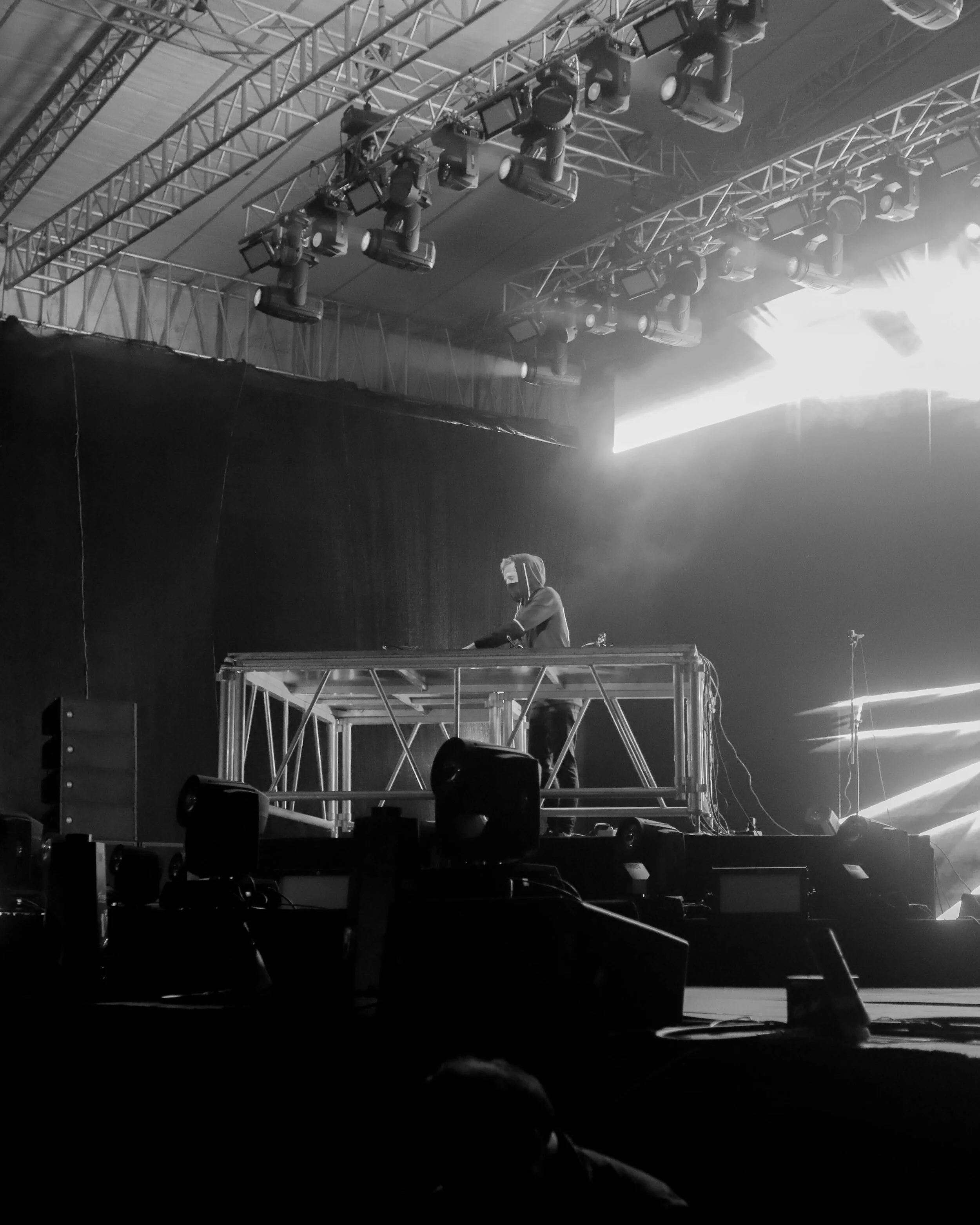 A DJ wearing a hoodie and headphones performing on a stage with lighting and sound equipment, with a black backdrop and stage trussing overhead.