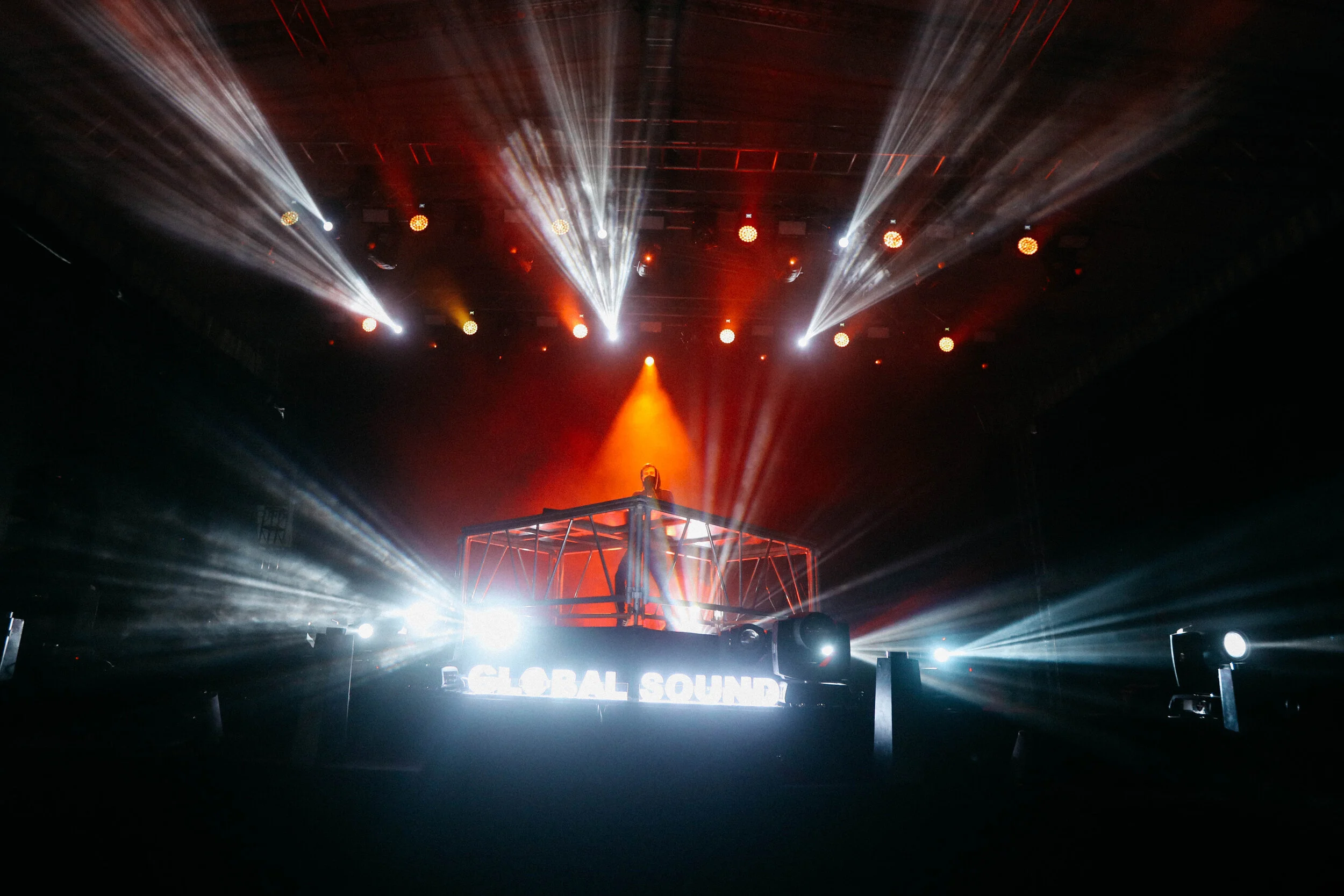 A DJ performs on a raised platform with bright red and white stage lights creating beams across a dark stage. The words 'GLOBAL SOUND' are illuminated in white on the front of the DJ booth.