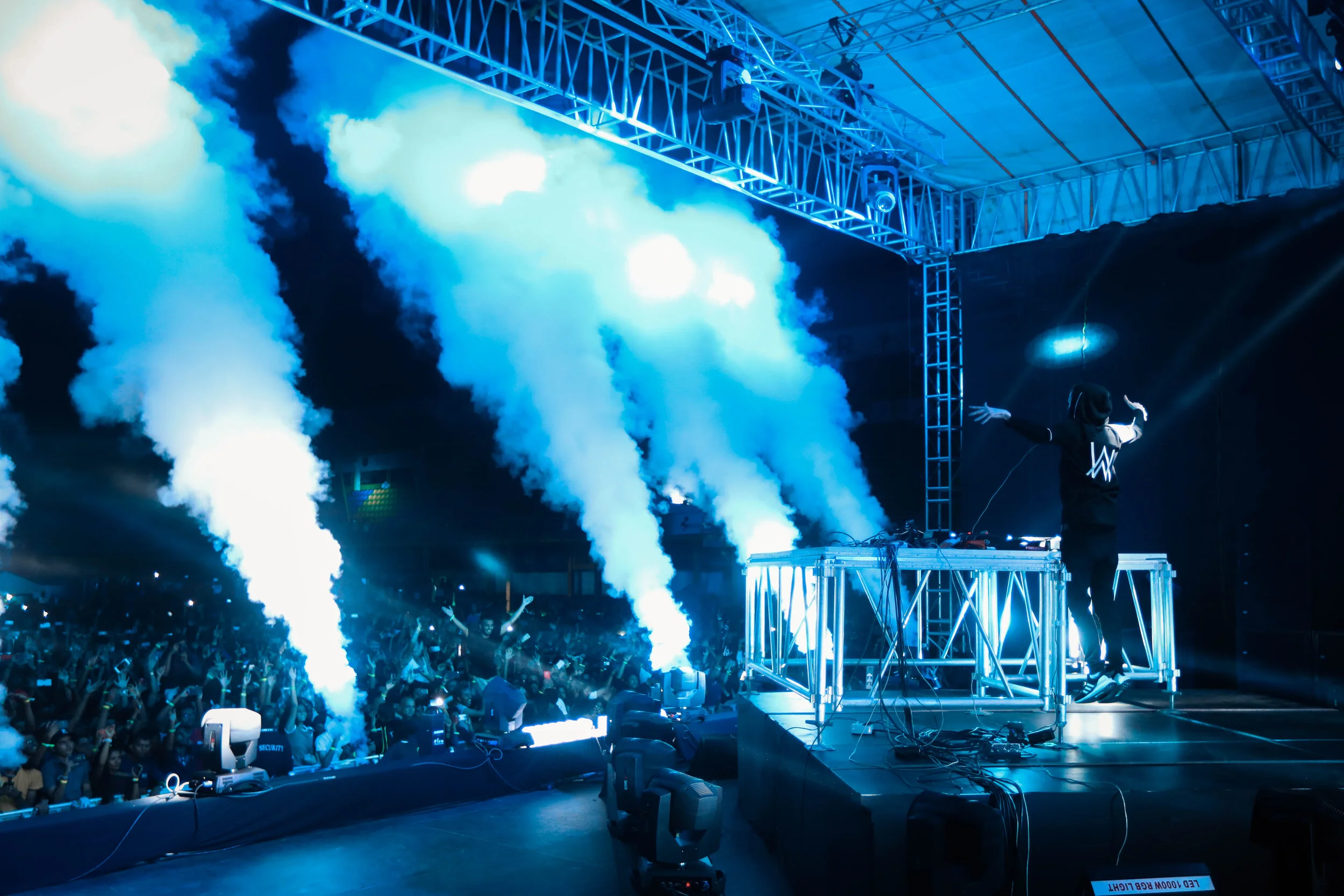DJ performing on stage with blue lighting and smoke effects, a large crowd dancing in front.