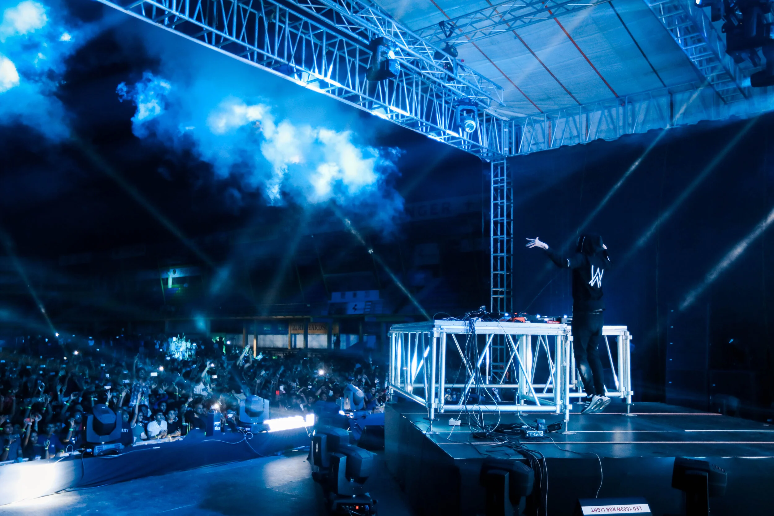 A DJ performs on an illuminated stage with blue lighting and smoke effects at a large indoor concert or music event with a crowd of people in the audience.