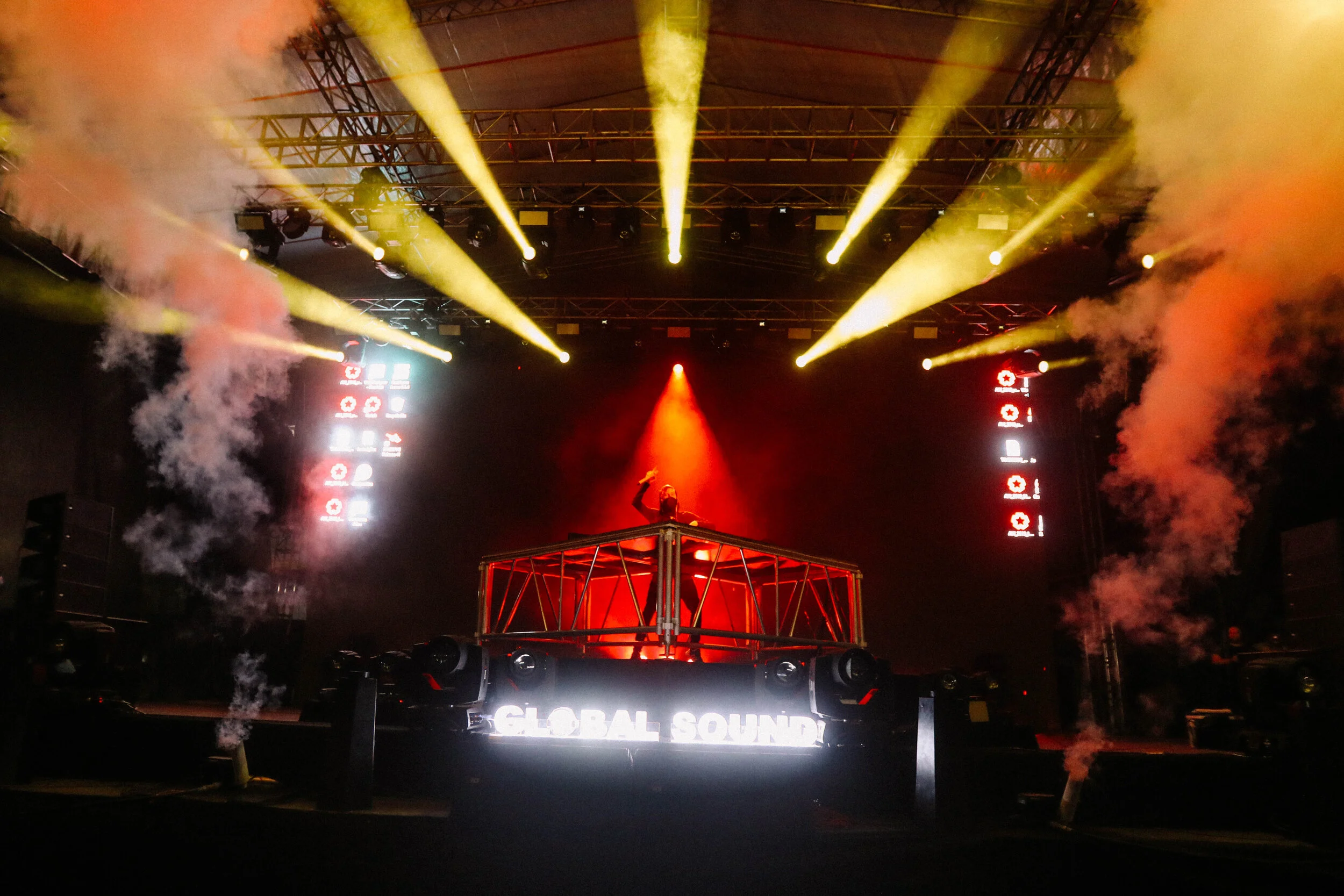 A DJ performs on stage with colorful lighting and smoke effects, standing behind a DJ booth labeled 'Global Sound' in a concert or club setting.