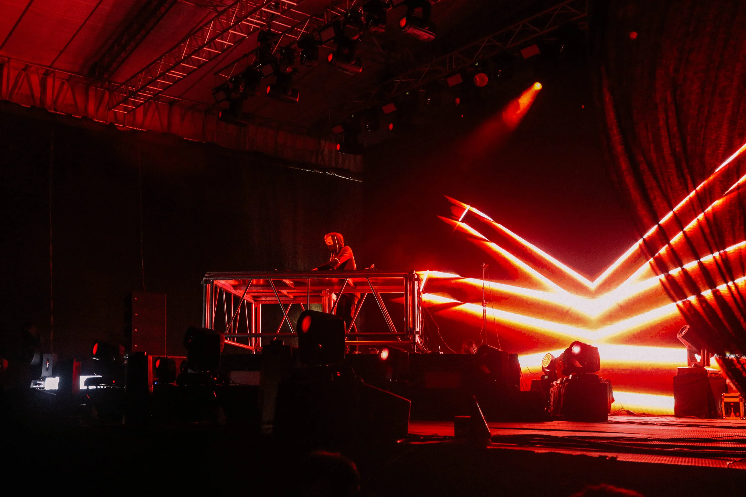 A DJ performing on stage with red and orange laser lights in the background, large speaker monitors, and stage lighting overhead.