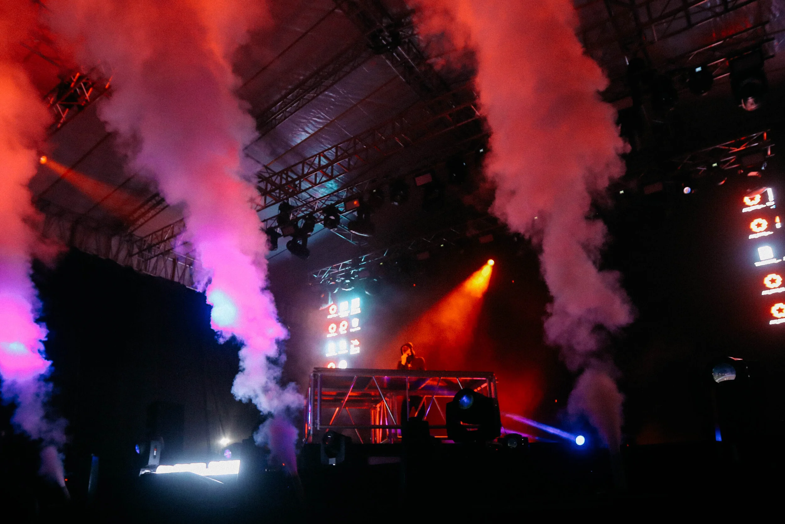 A DJ performing on an elevated stage with colorful smoke effects and LED screens in the background, during a concert or music event.