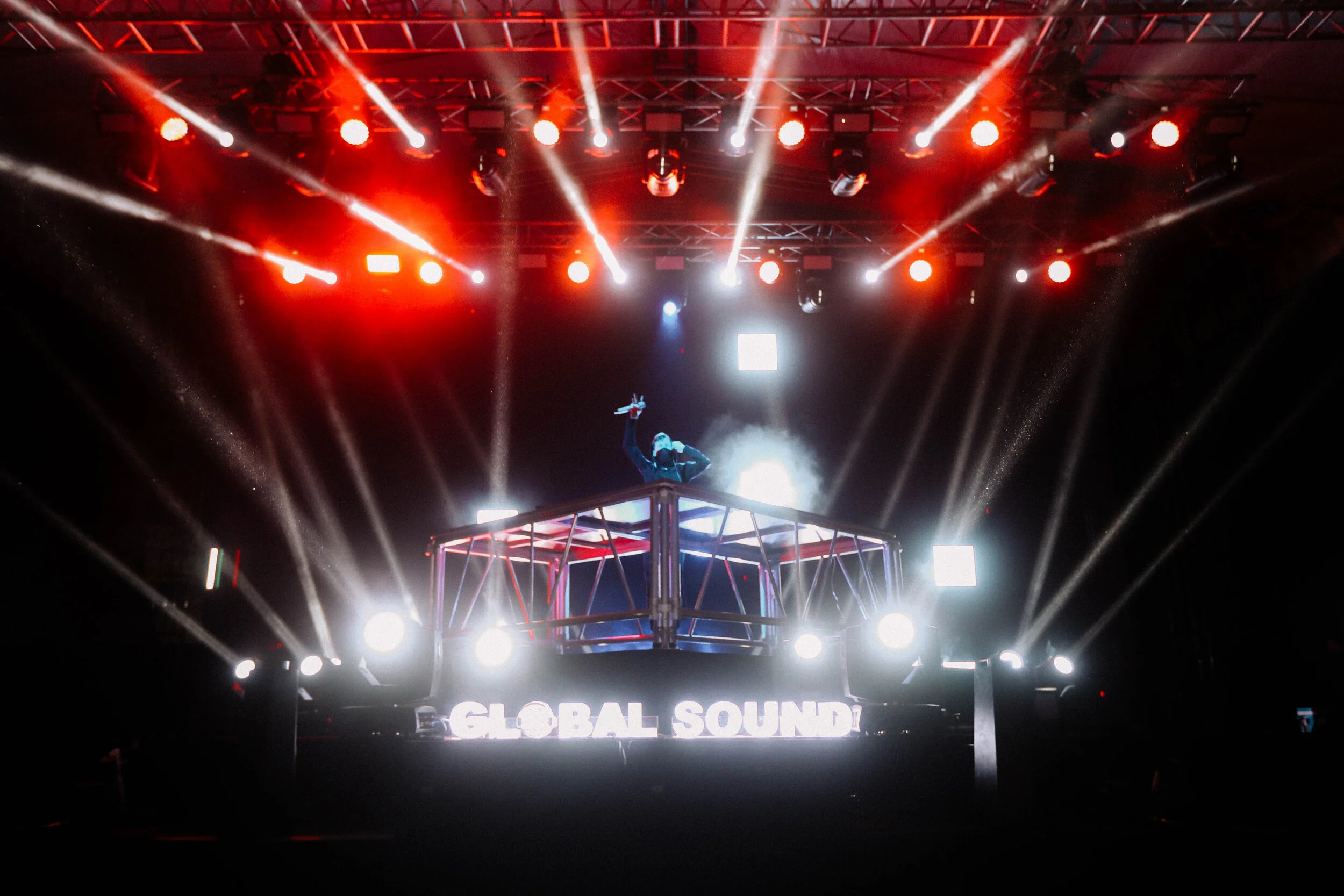 A DJ performs on an elevated stage with bright red, white, and blue lights shining down, and a large screen displaying the words "GLOBAL SOUND".