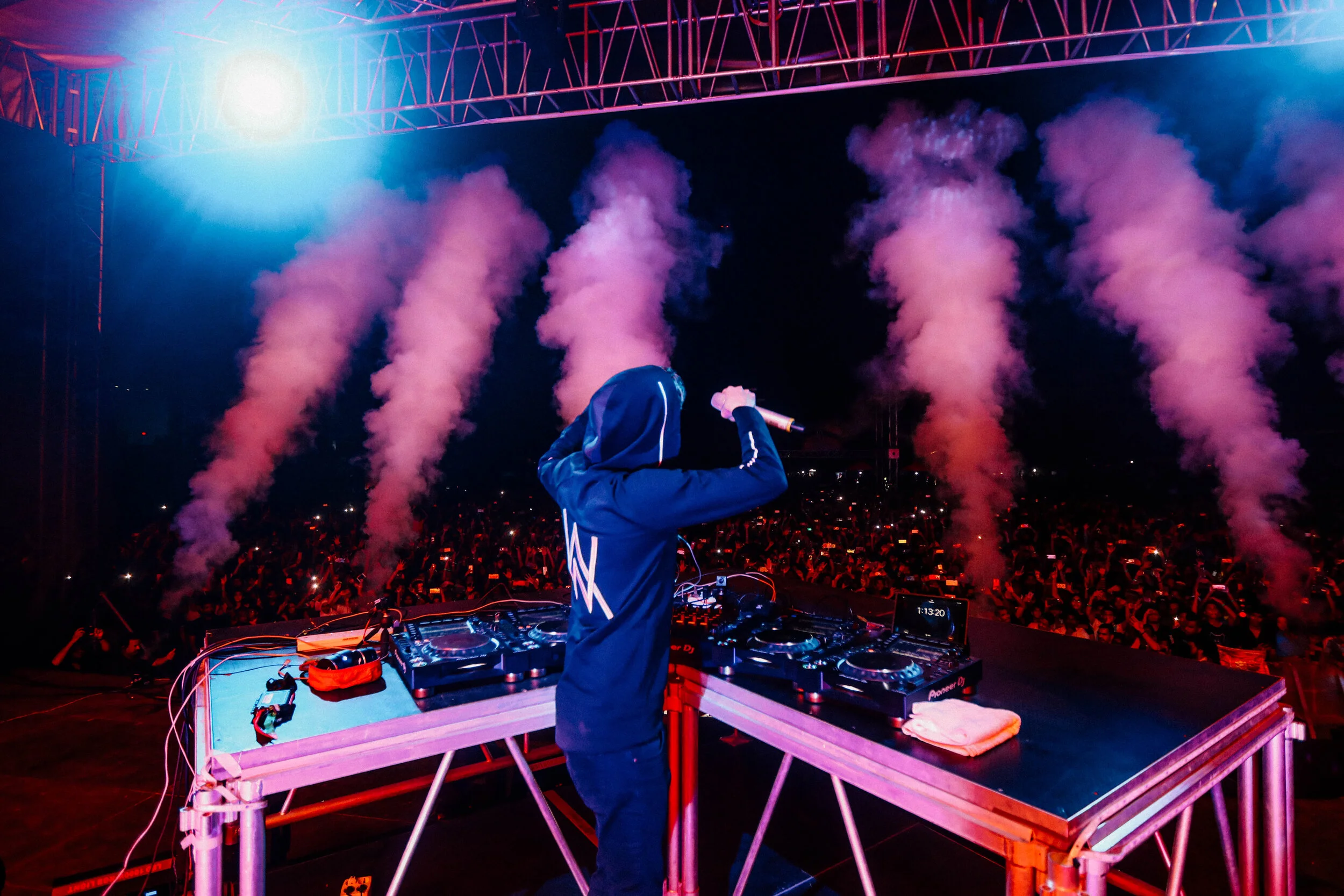 A DJ wearing a hoodie performs at a nighttime outdoor concert with pink smoke and bright lights, facing a large crowd.