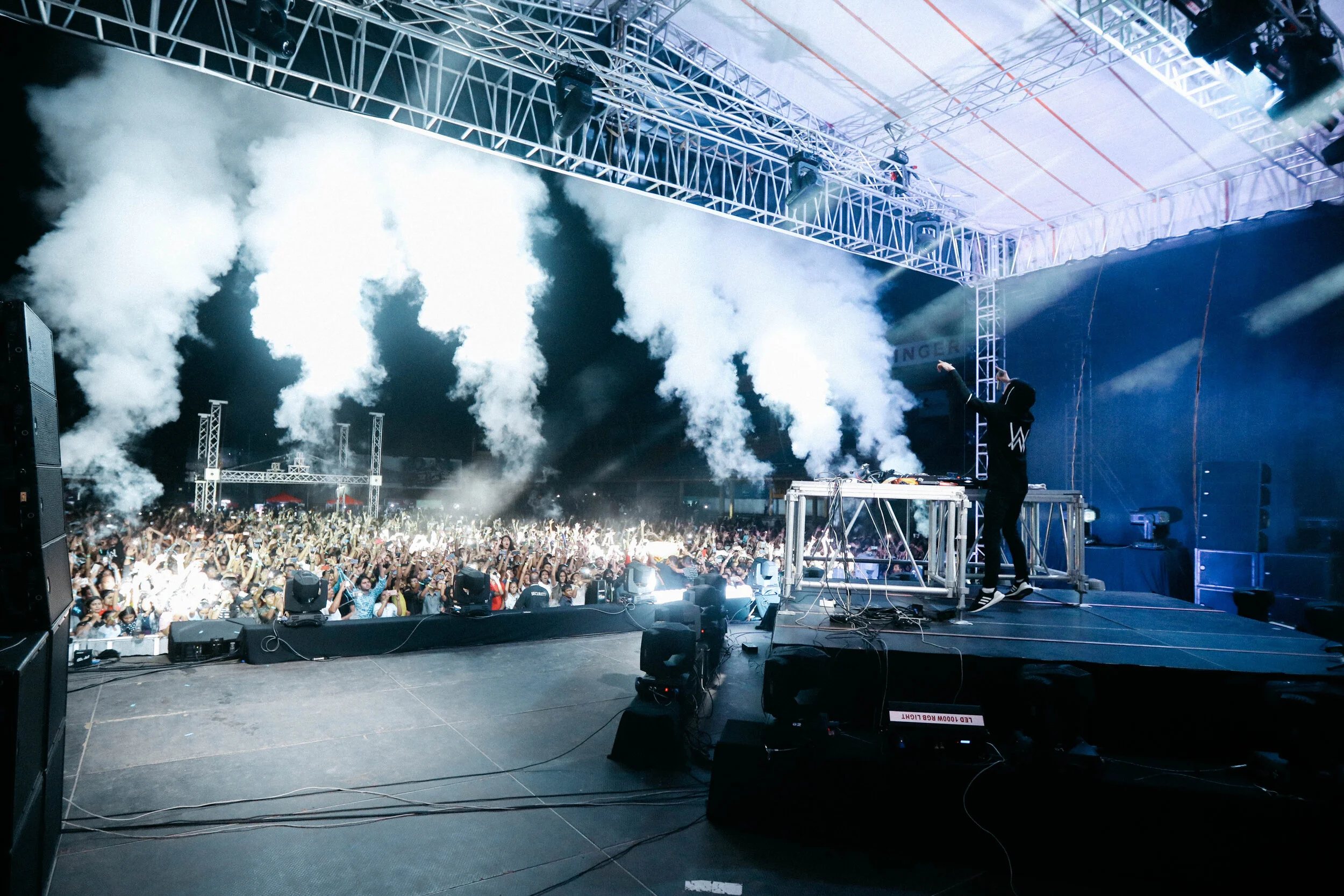 A DJ performs on stage at a music festival, with crowd and smoke effects, large speakers, and lighting rig overhead.