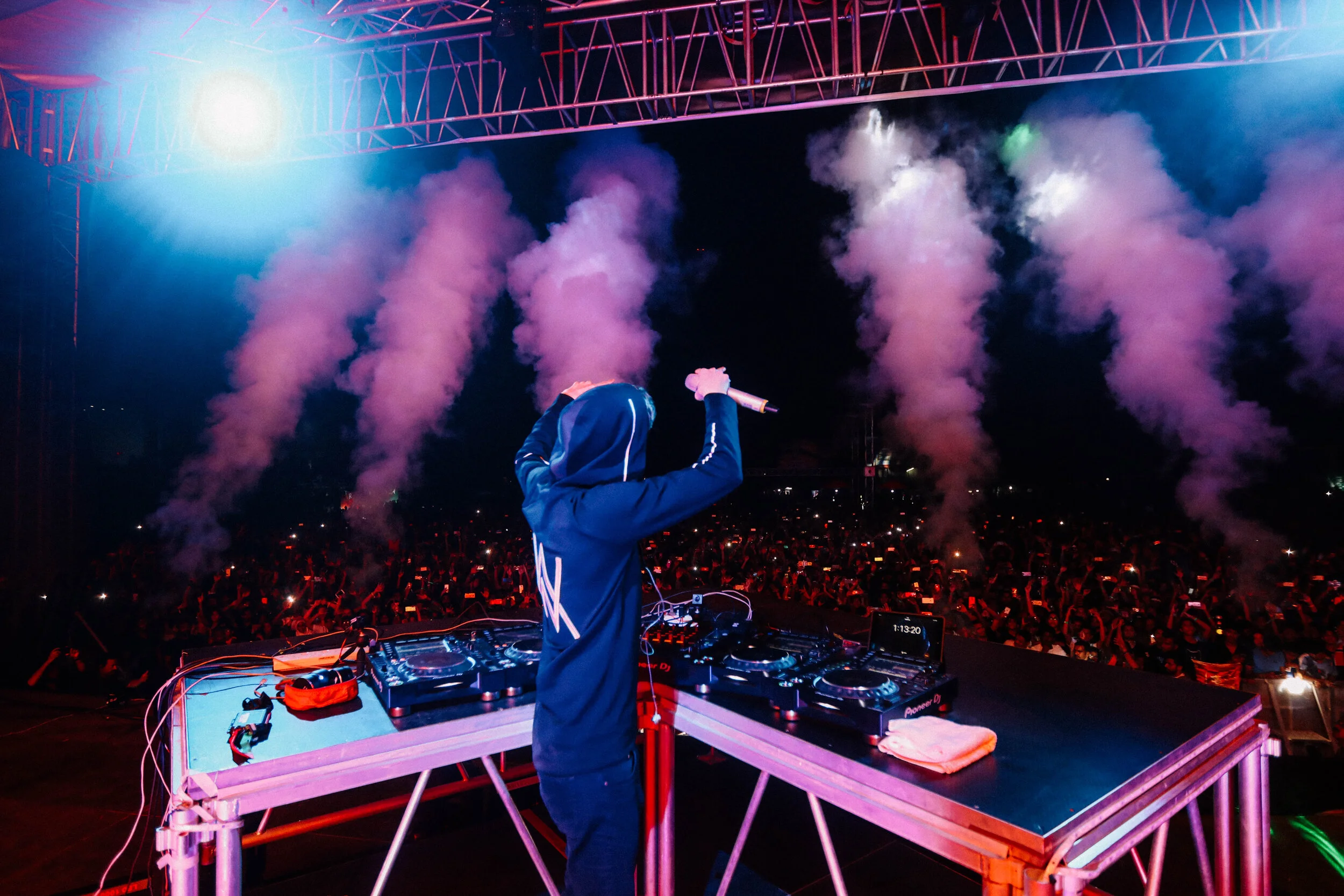 A DJ performs on stage at a concert during the night, surrounded by smoke and colorful lights, with a large crowd in front wearing glow accessories.