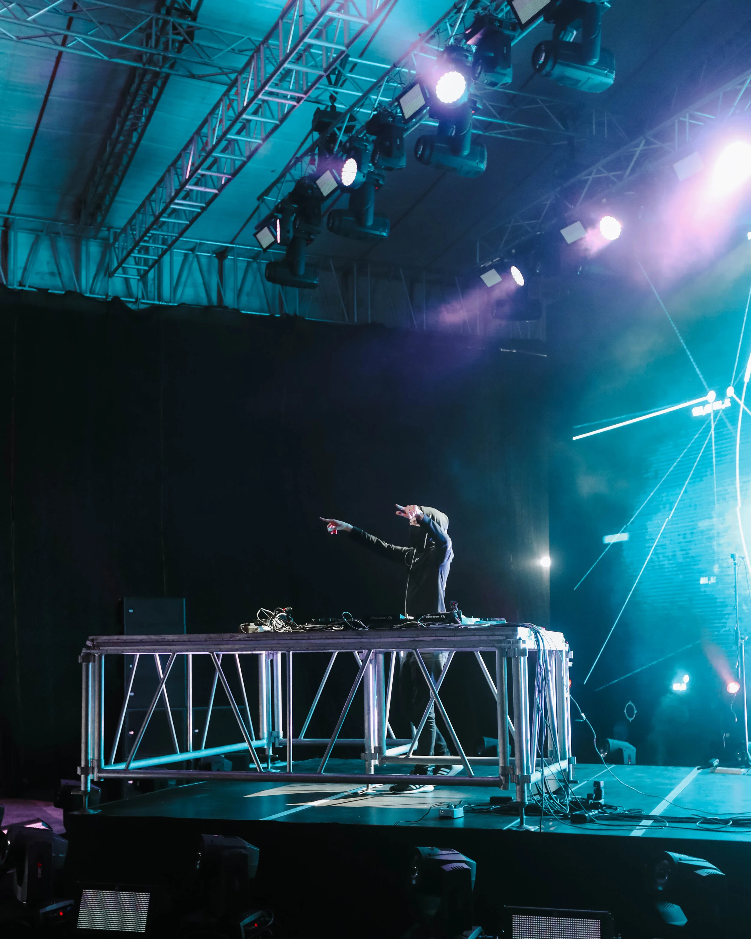 DJ performing at a concert on a stage with colorful lighting and rigging overhead.