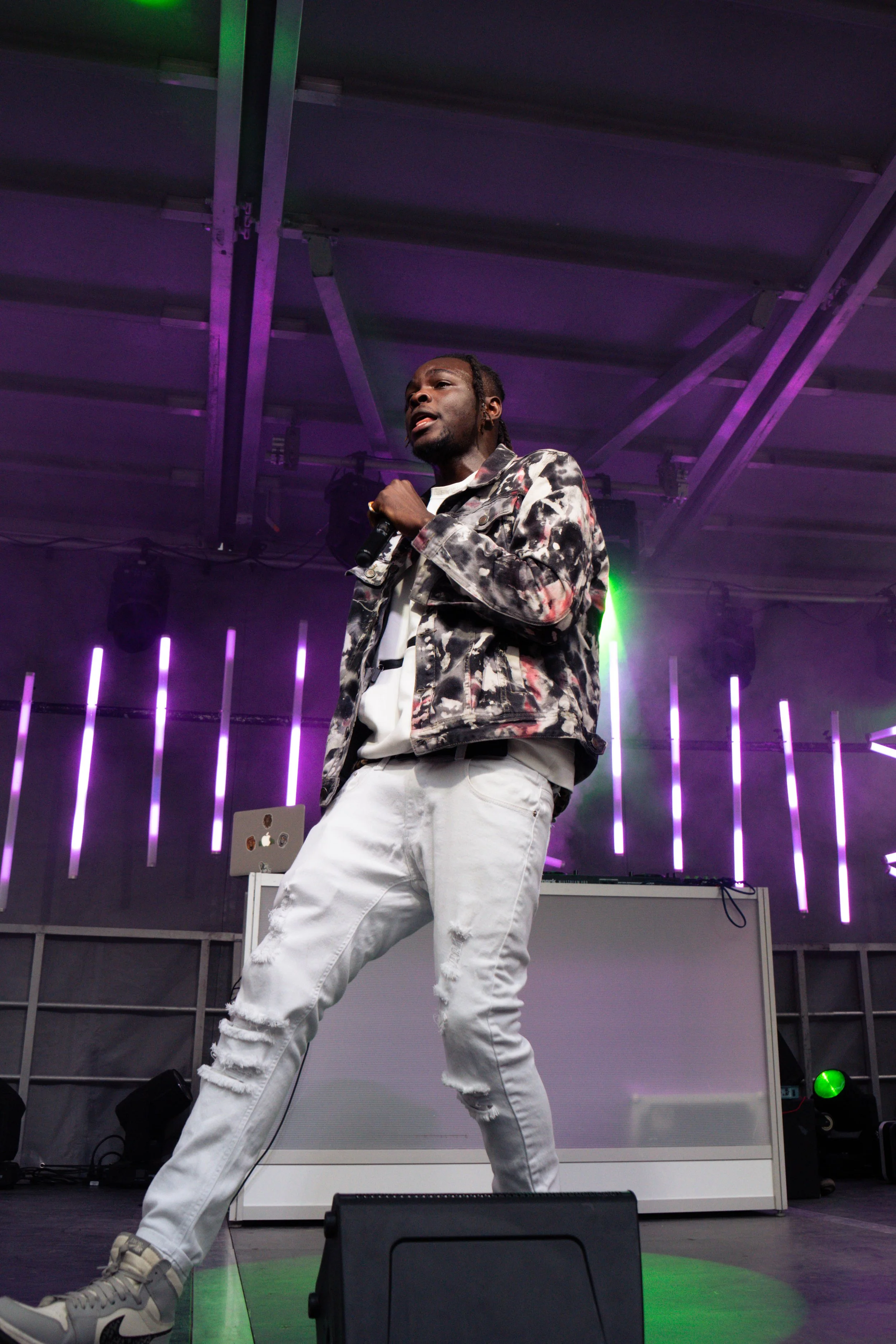 A male performer singing into a microphone on stage with purple and green lighting, wearing a patterned jacket and distressed white jeans.