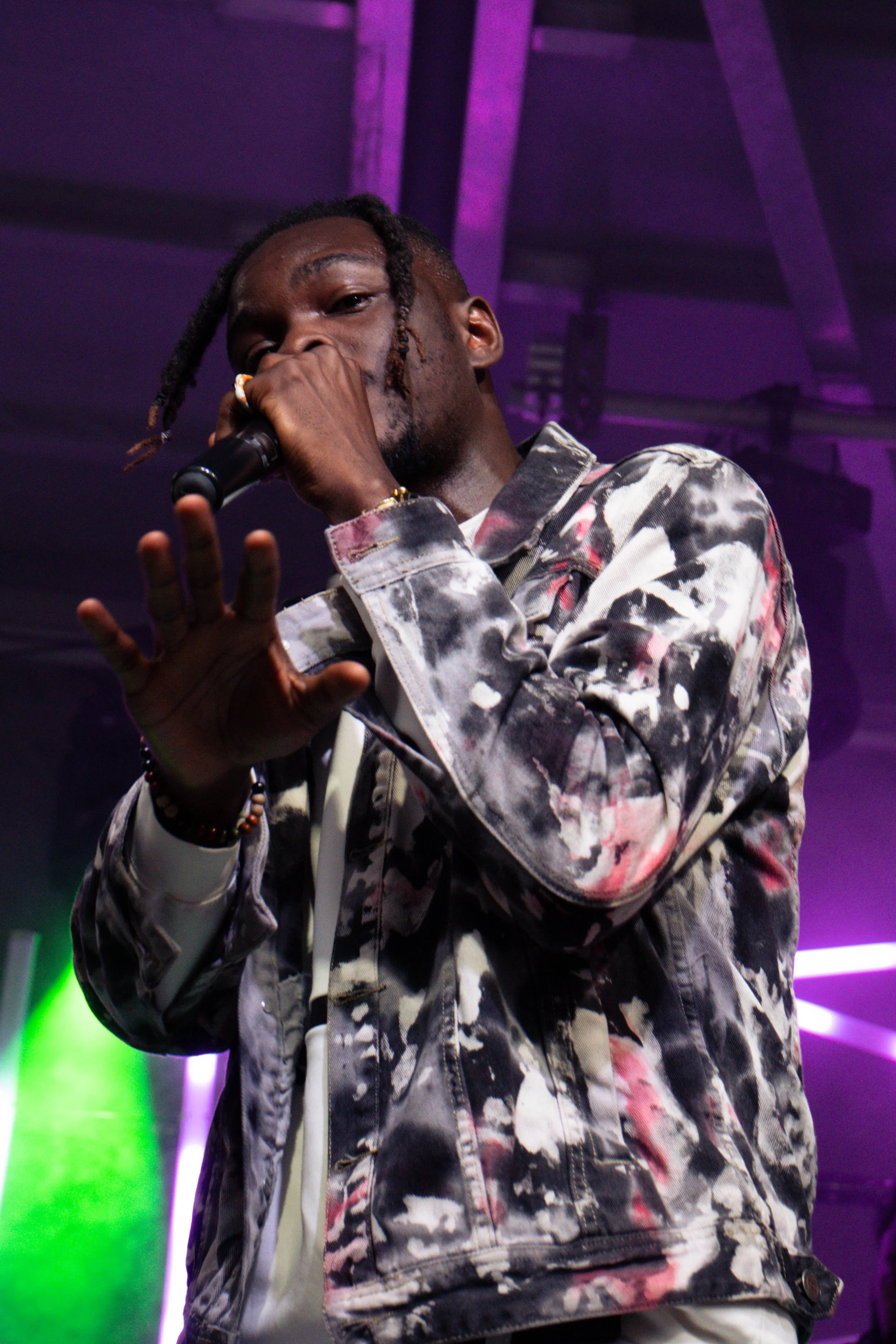 A man with dreadlocks and a tattooed jacket holding a microphone, performing on stage with purple and green lighting in the background.