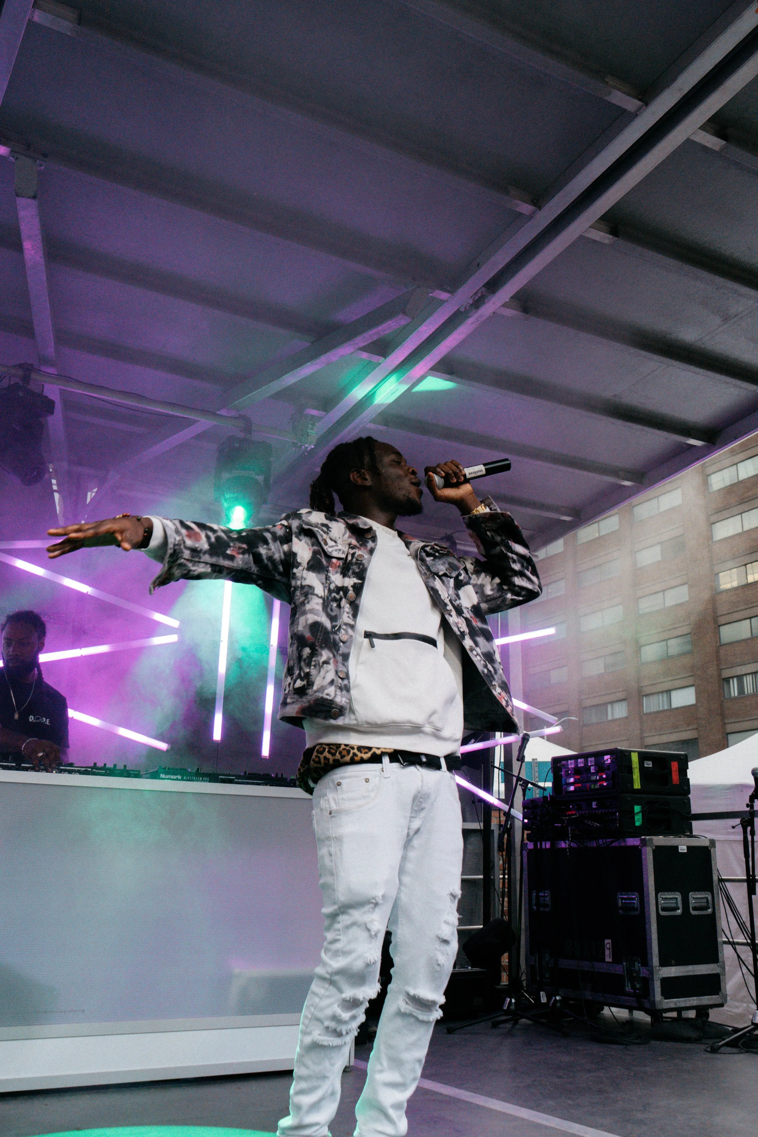 A male performer singing into a microphone on stage with purple and green lighting and electronic equipment in the background.