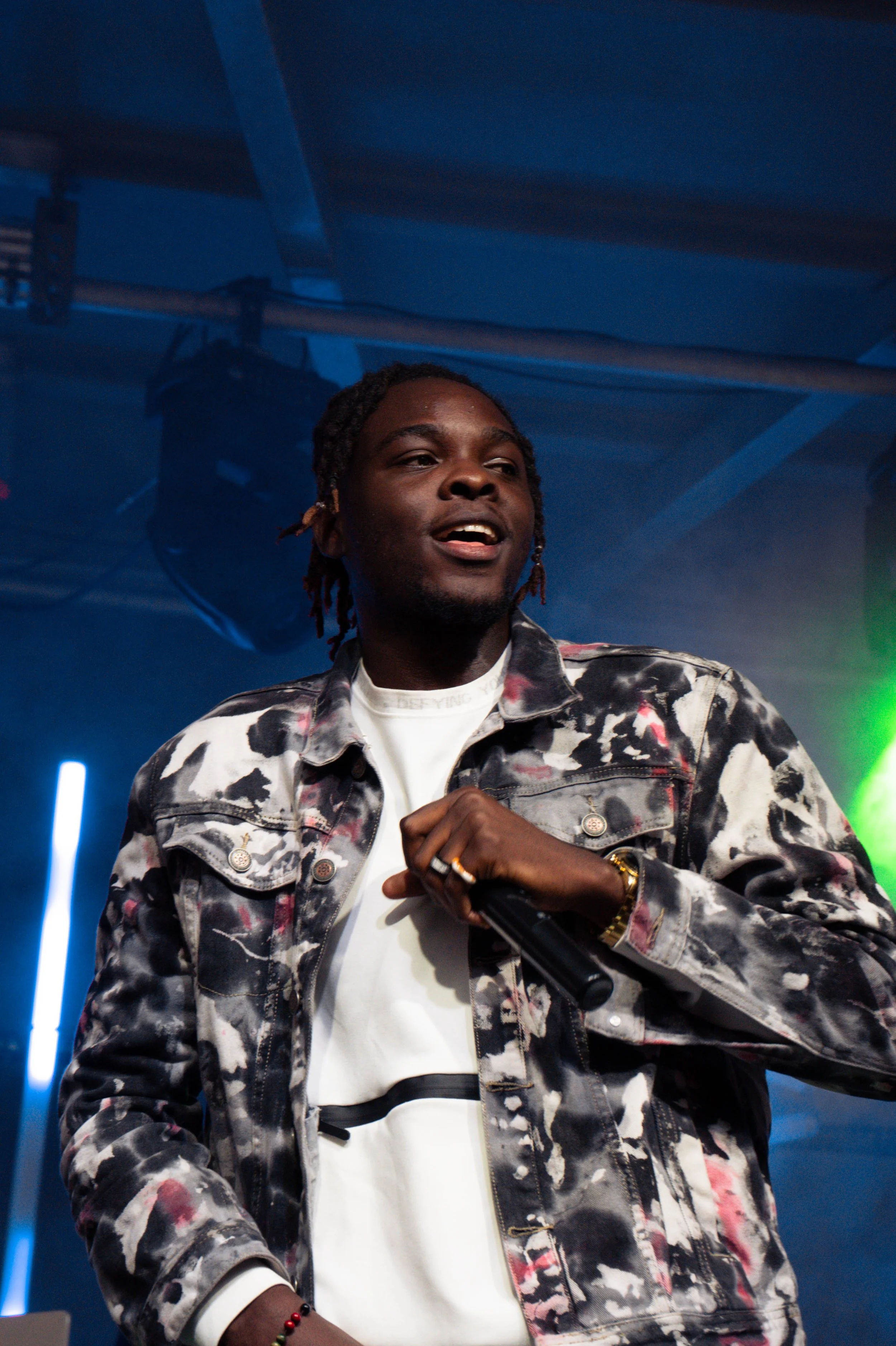 A young man with dreadlocks in a patterned jacket holding a microphone on a stage with colored lights.