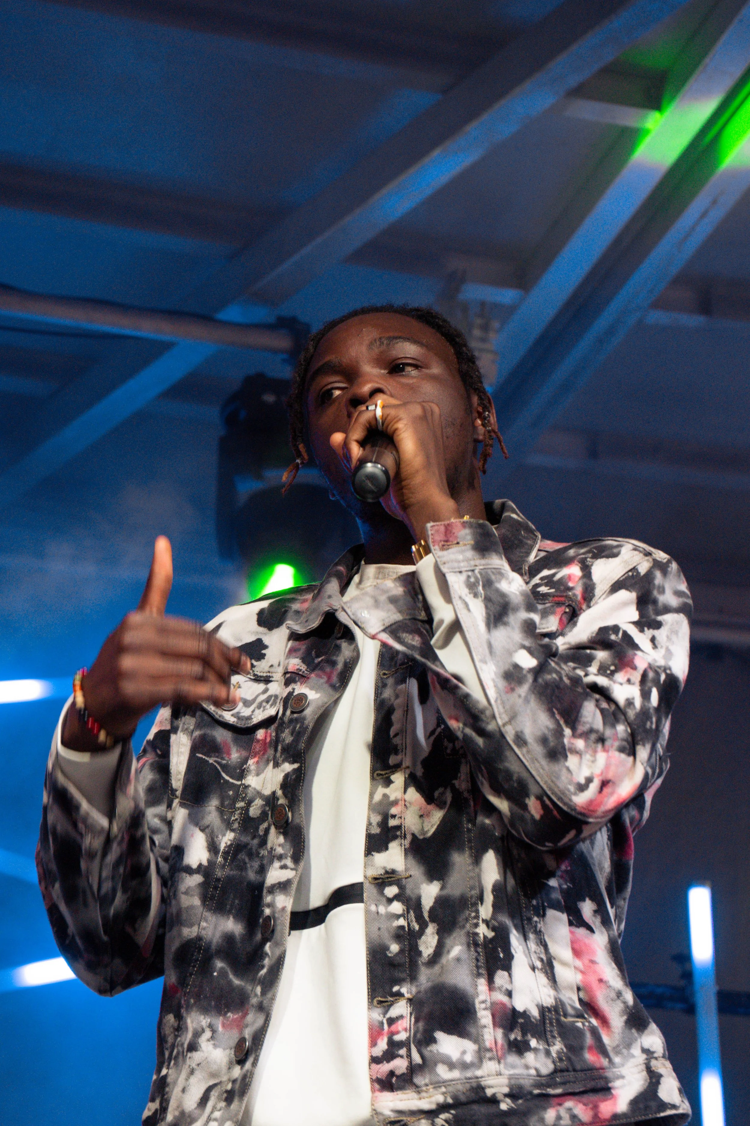 Hip-hop artist performing on stage with microphone, wearing a patterned denim jacket, in a setting with colorful stage lighting.