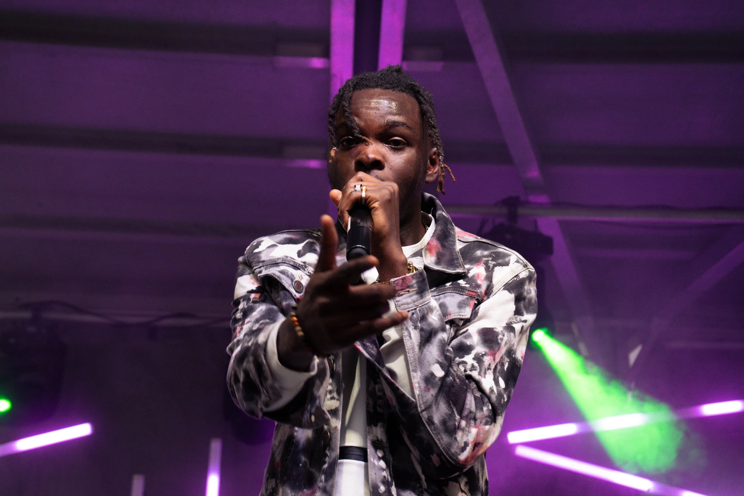A man with dreadlocks singing into a microphone on stage, with purple and green stage lighting.
