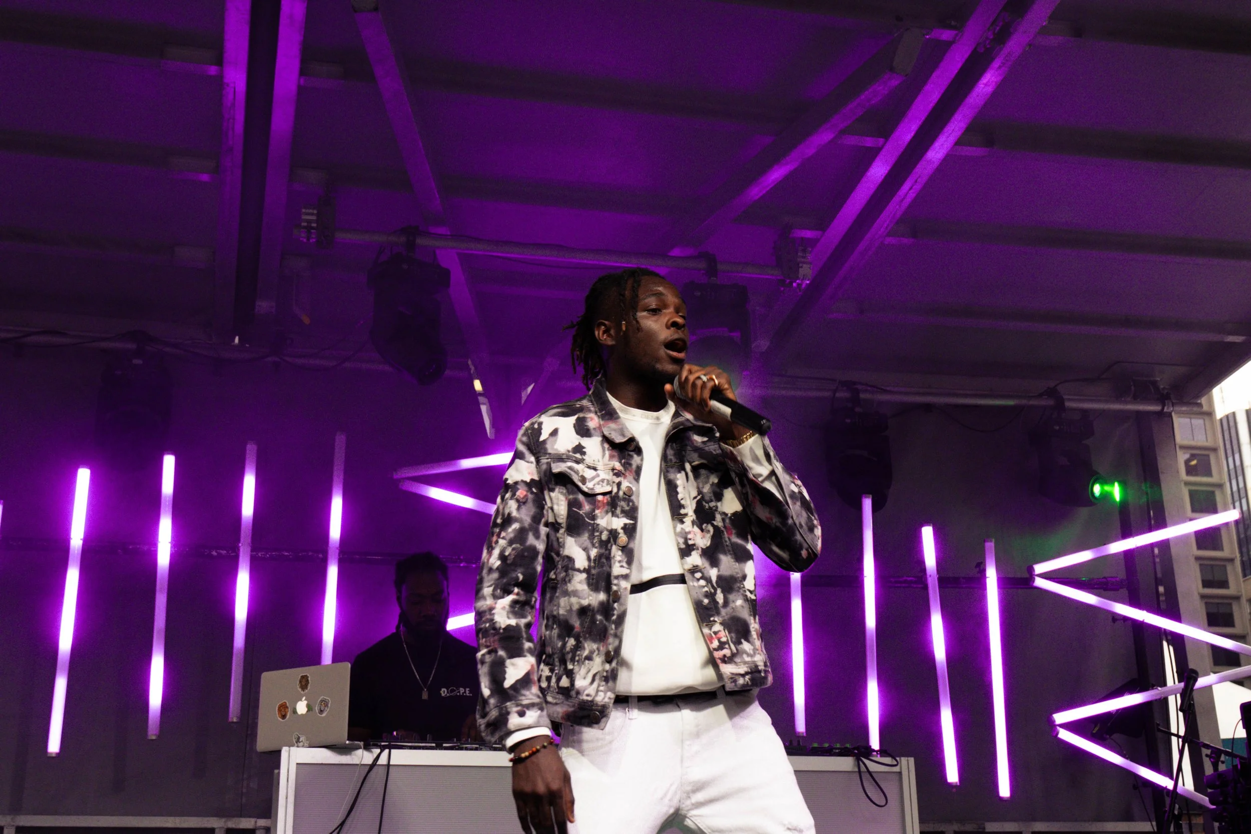 A male performer singing into a microphone on a stage with purple neon lights in the background, a DJ working behind him, and city buildings visible outside.