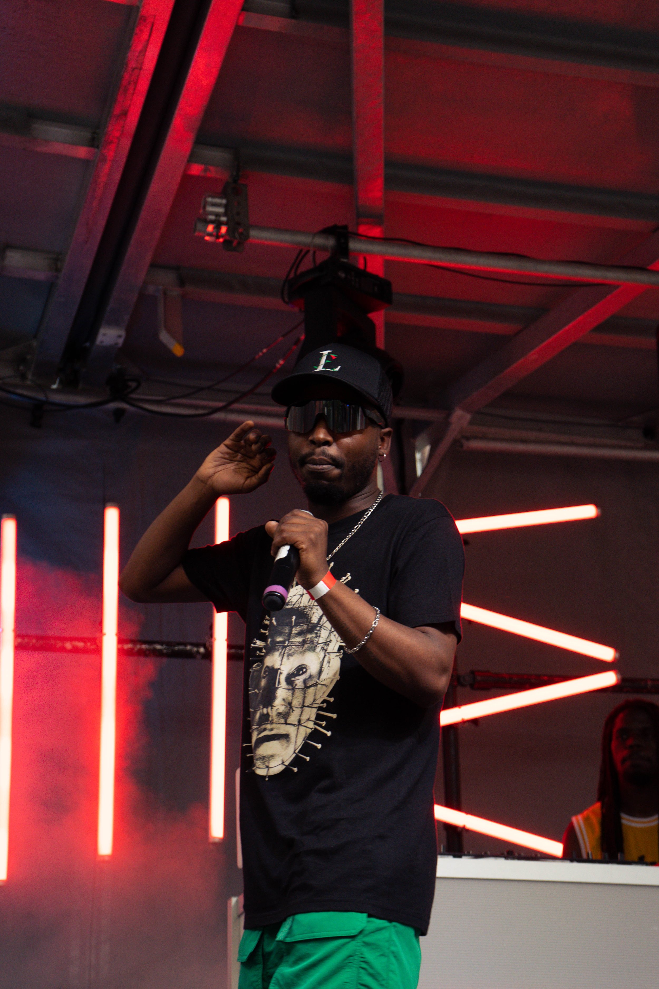 A man wearing a black baseball cap, sunglasses, a black T-shirt with a graphic face design, and green shorts, holding a microphone on stage with red LED lights in the background.