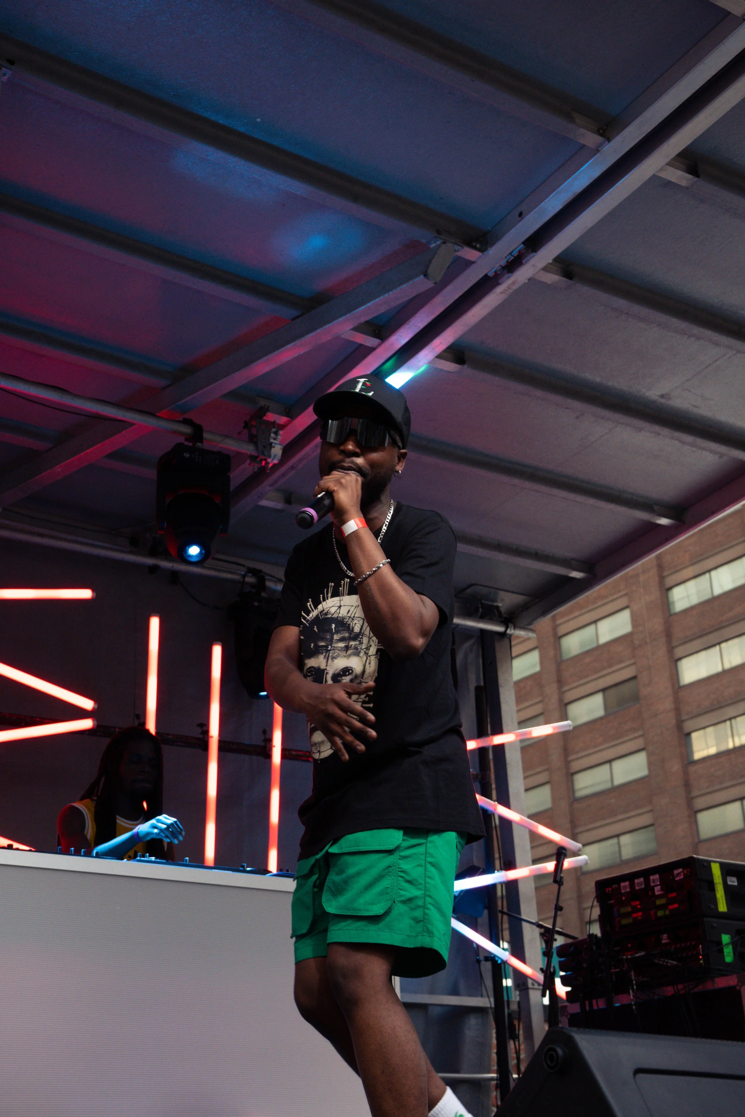 A man in sunglasses, a cap, and casual clothing holds a microphone on a stage with a DJ behind him, illuminated by colorful stage lights during a live performance in an outdoor urban setting.