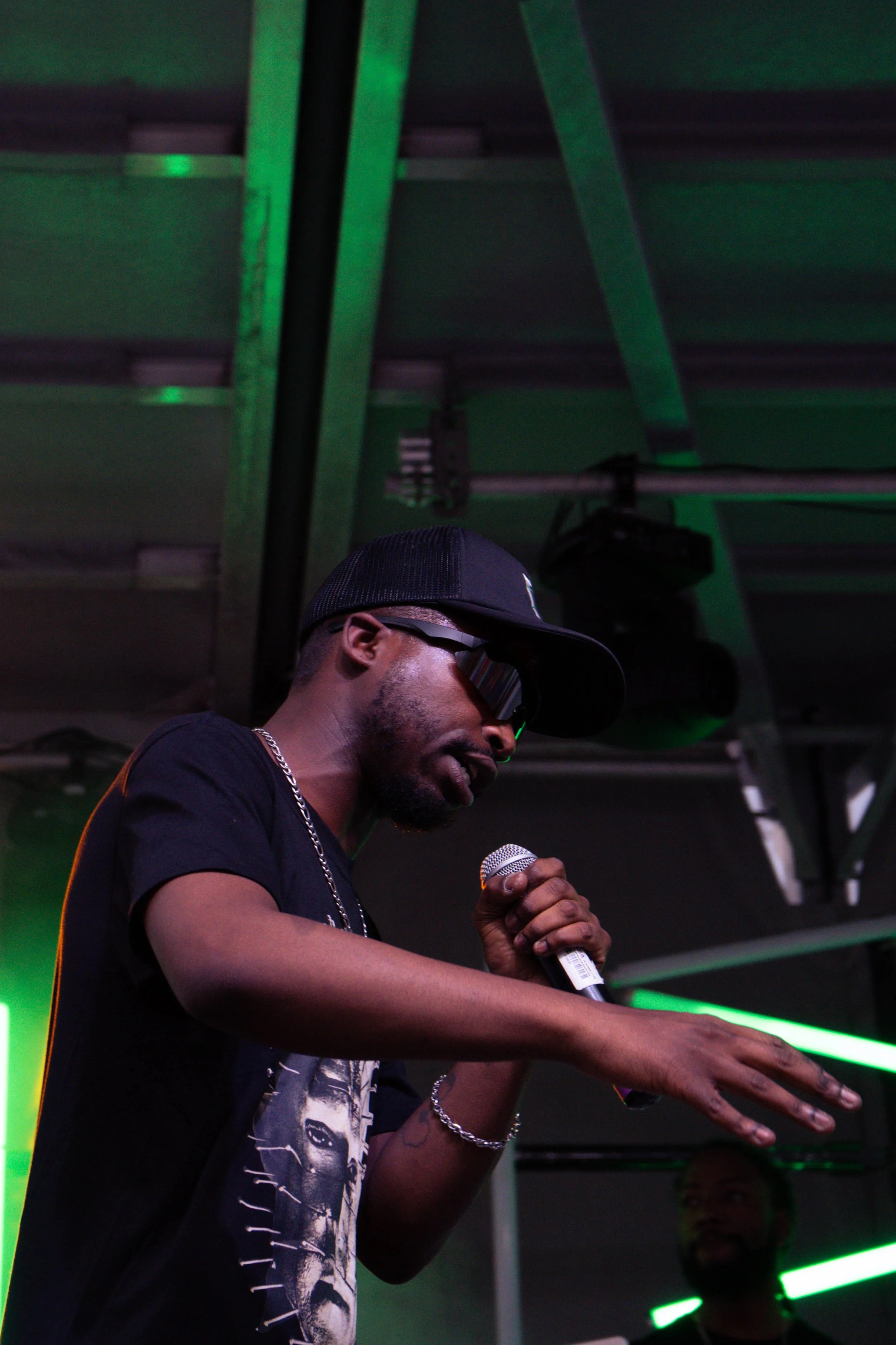 A man wearing sunglasses, a black baseball cap, a black T-shirt with a graphic print, and jewelry, is holding a microphone and speaking or singing at an event with green lighting.