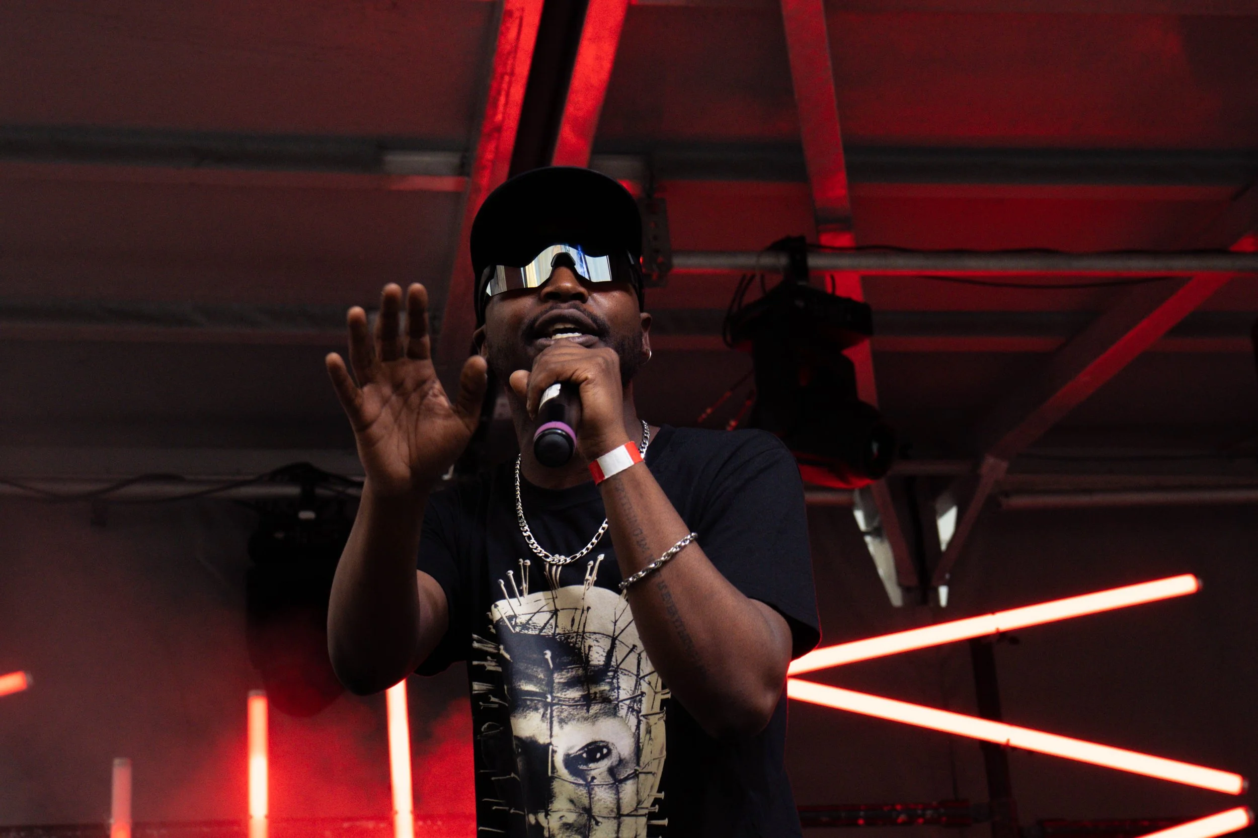 A man wearing sunglasses, a black cap, and a black T-shirt with a lion graphic, holding a microphone and speaking on stage with red neon lights in the background.