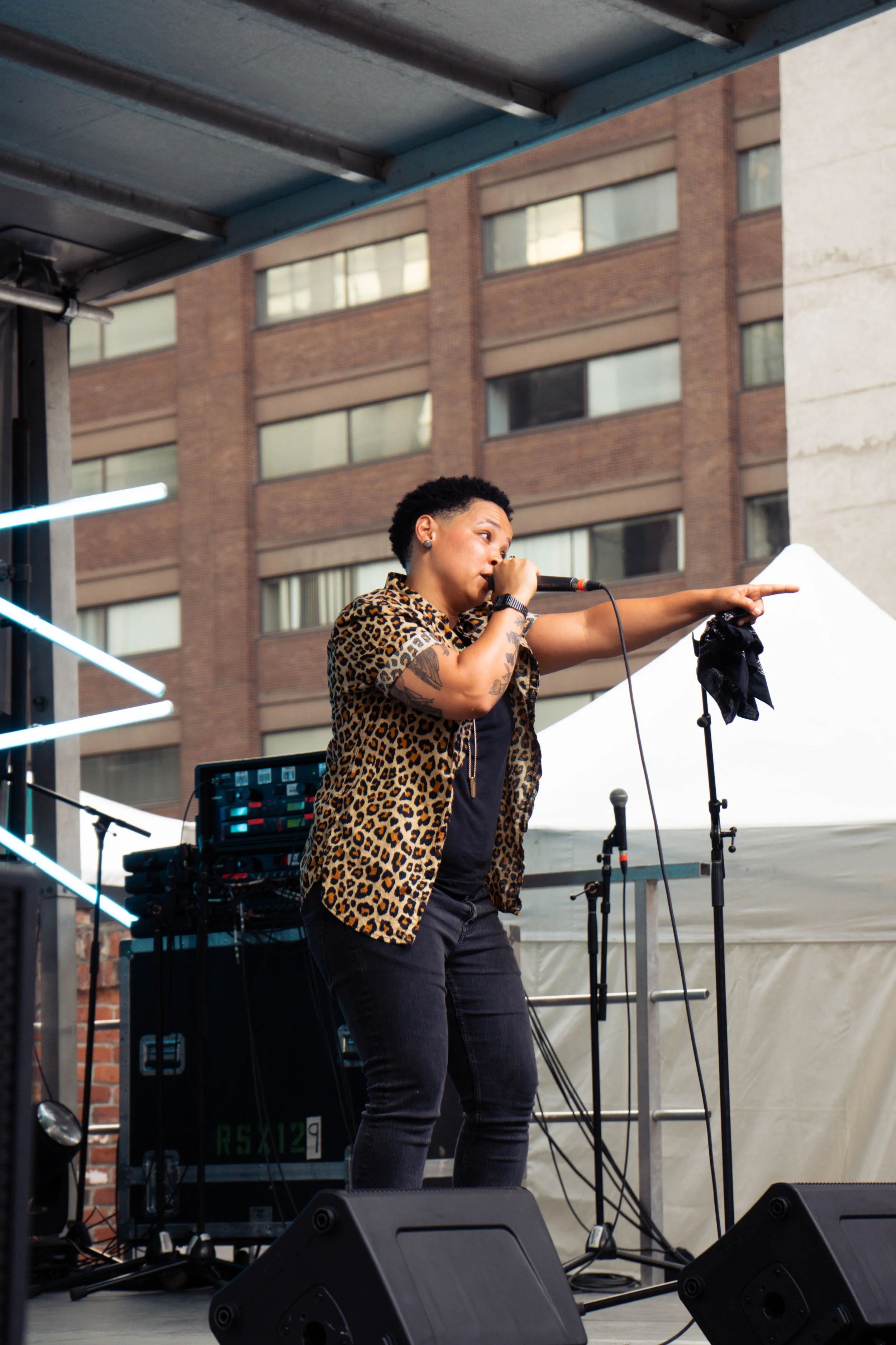 A woman with short curly hair, wearing a leopard print shirt, singing into a microphone on an outdoor stage with a city building in the background.