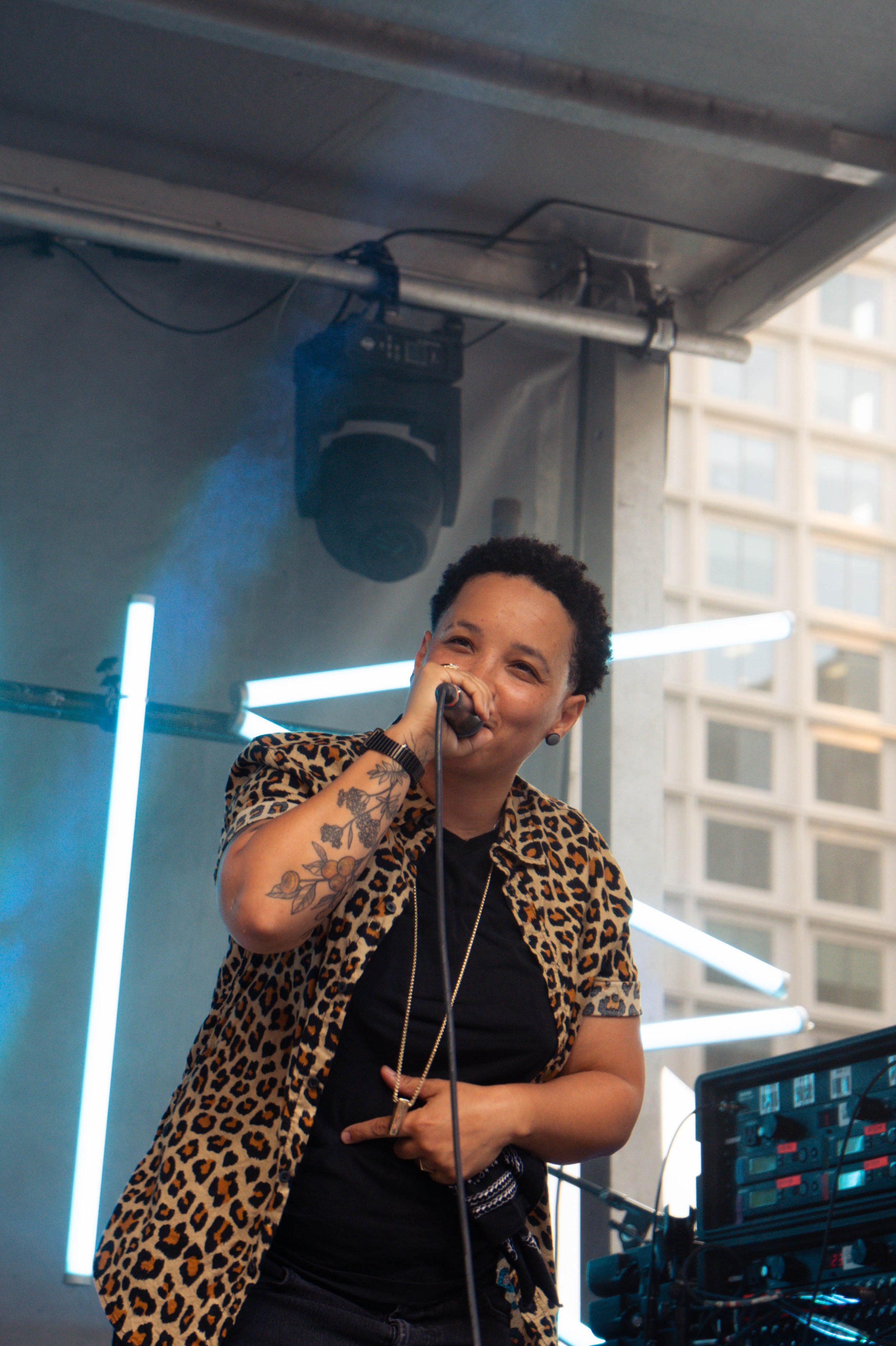 A woman with short curly hair, tattoos on her arm, and a leopard print shirt, singing into a microphone at a music event with neon lights and electronic equipment around her.