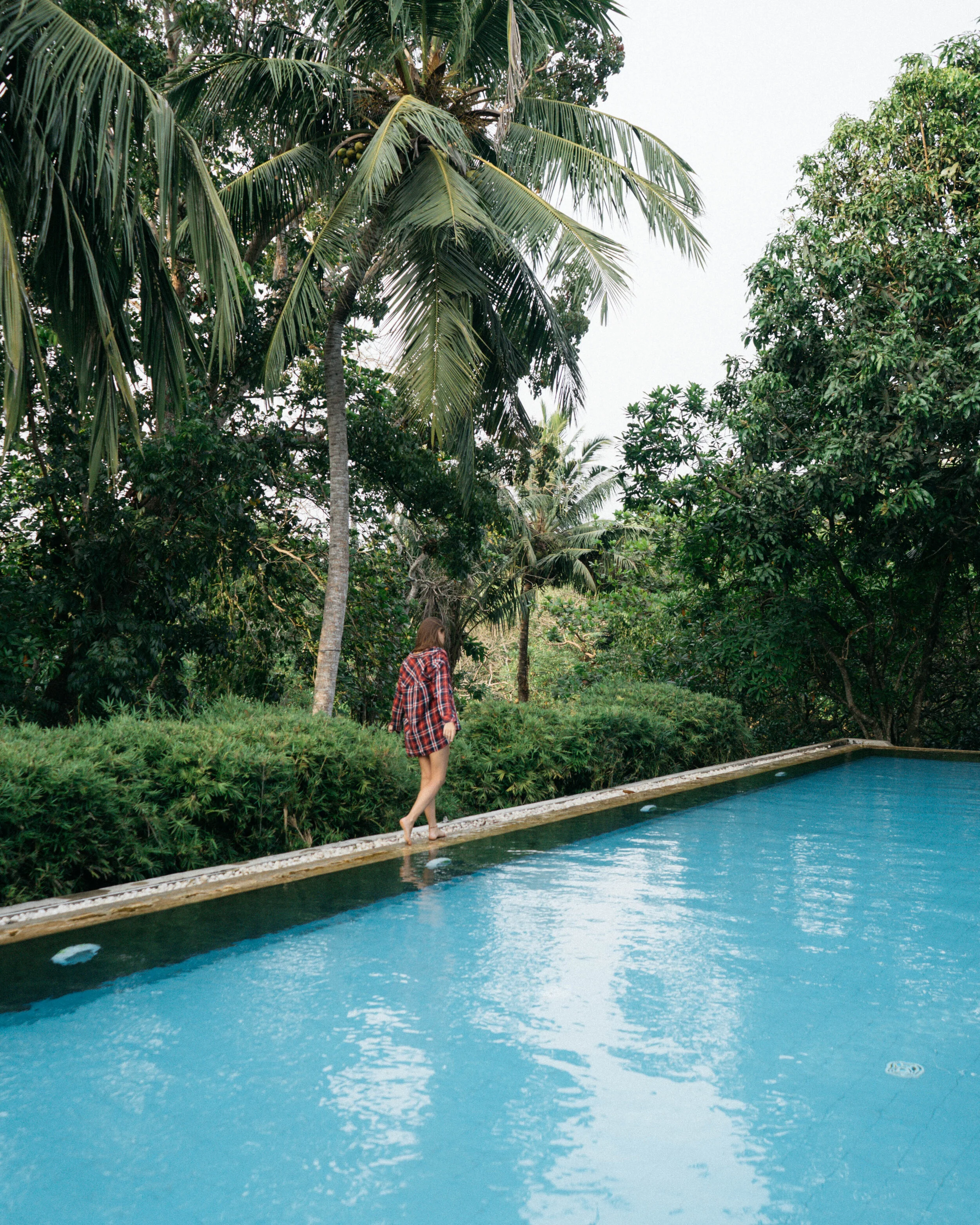 A person in a red plaid shirt walks along the edge of a swimming pool surrounded by lush green tropical trees