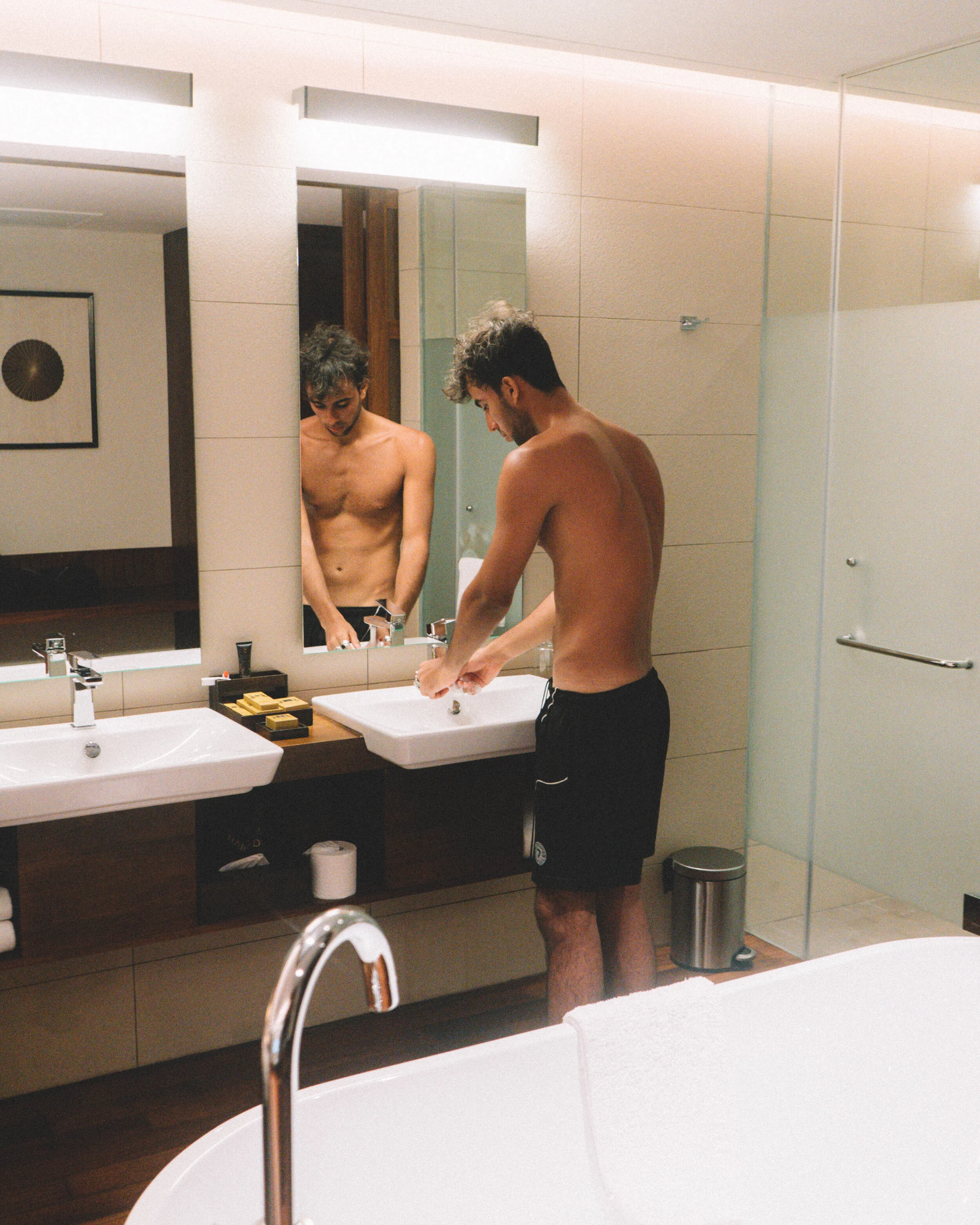 A shirtless man with curly hair and a beard washing his hands at a bathroom sink with a large mirror reflecting his image. The bathroom has beige tiled walls, a wooden vanity, and various toiletries on the counter.
