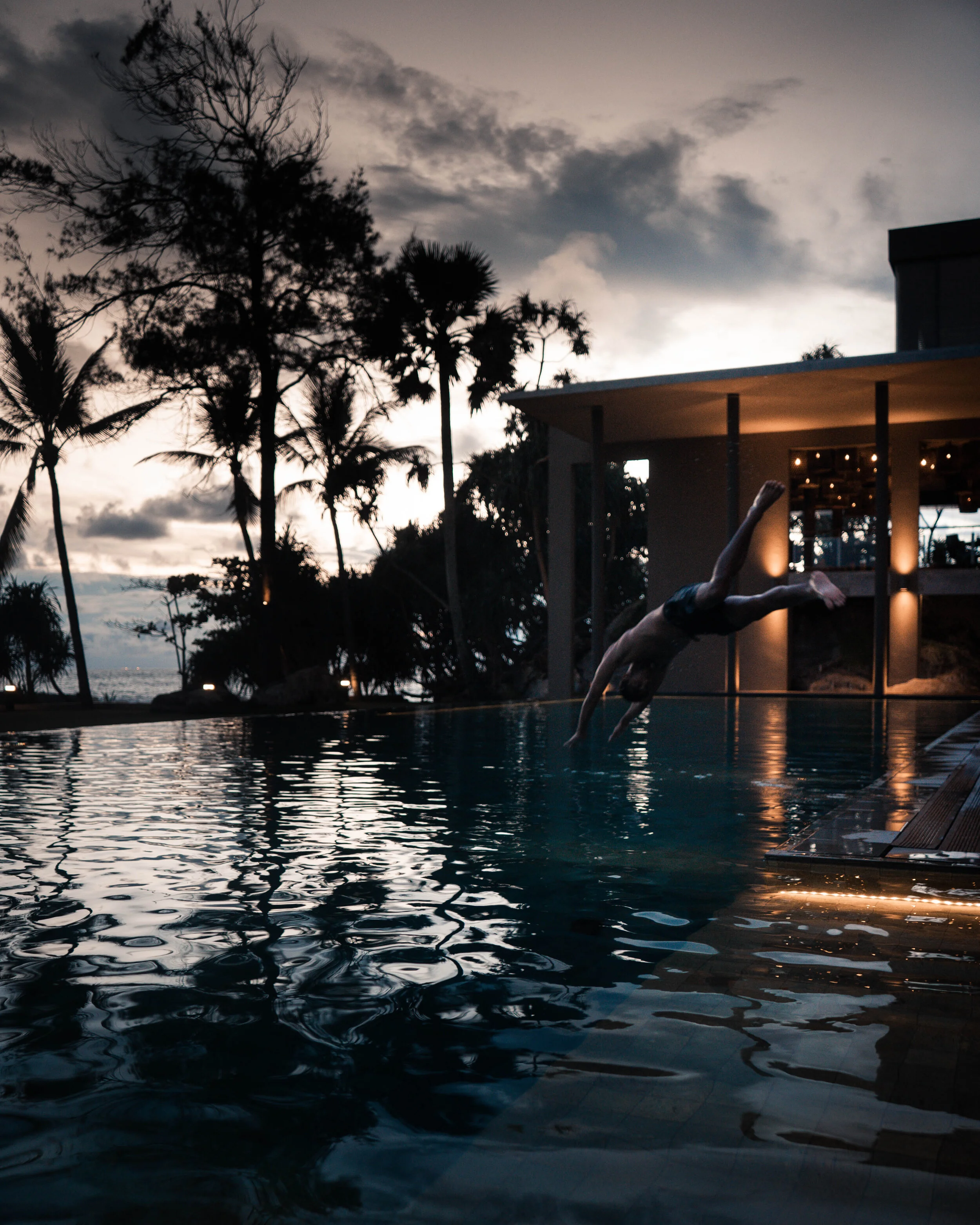 Person diving into a swimming pool during sunset with trees and a modern building in the background.