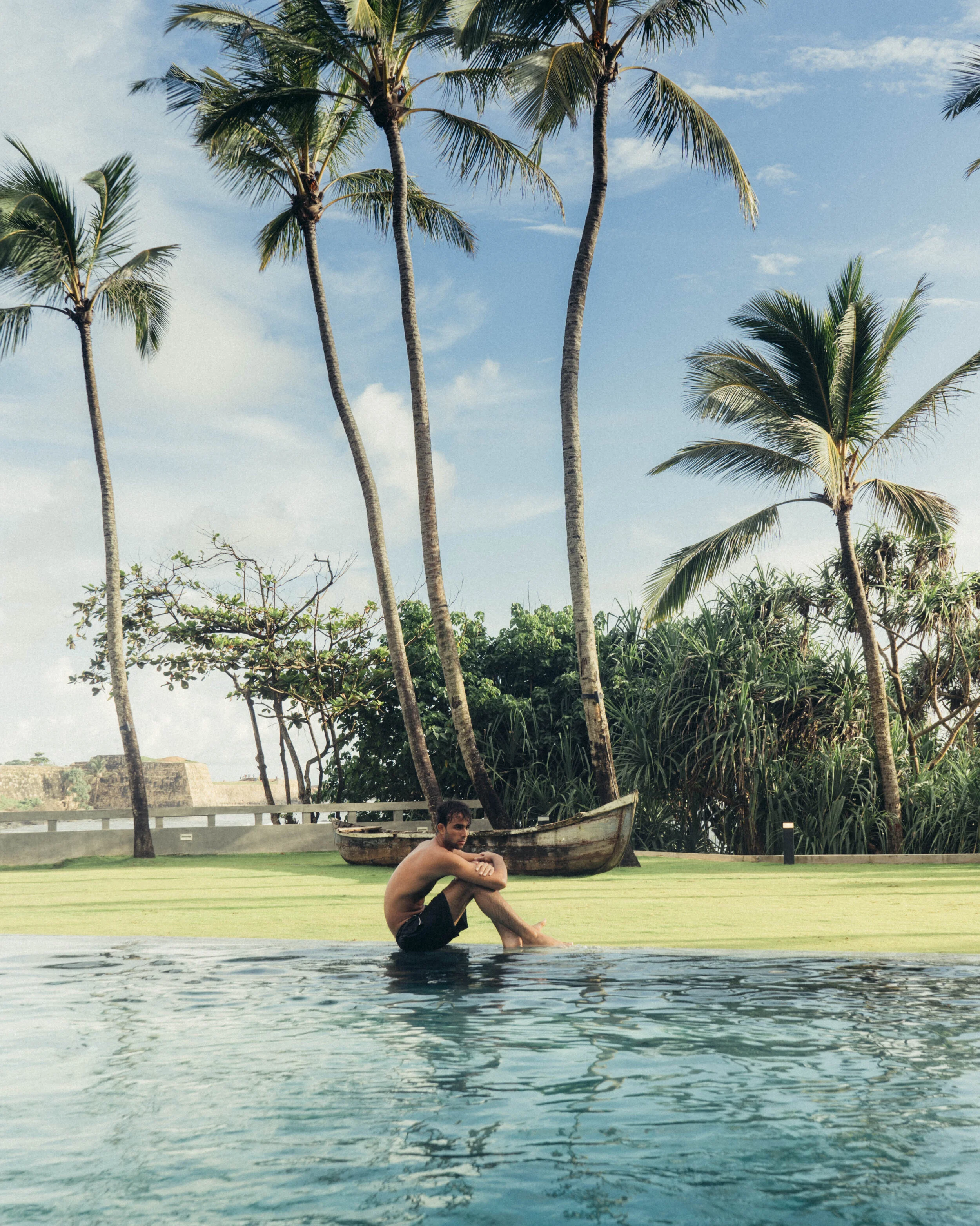 A man sitting by a swimming pool with a tropical background of palm trees, boat, and lush greenery on a sunny day.