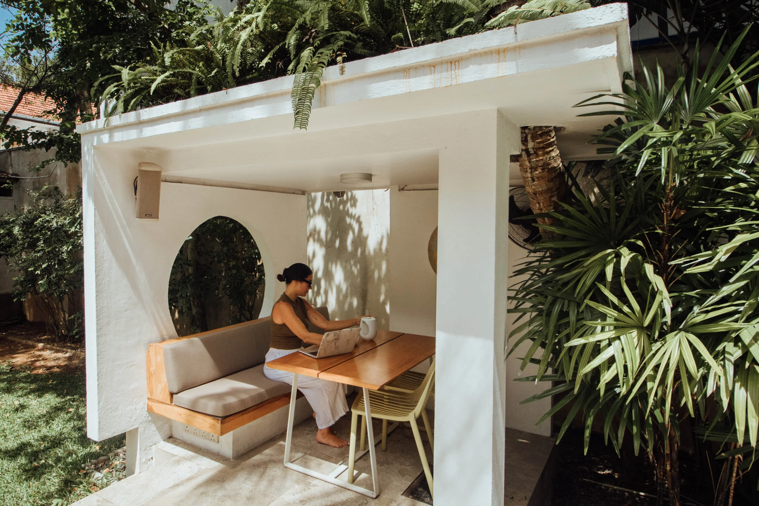 Woman sitting at an outdoor workspace with a laptop and coffee mug inside a small white pavilion surrounded by greenery.
