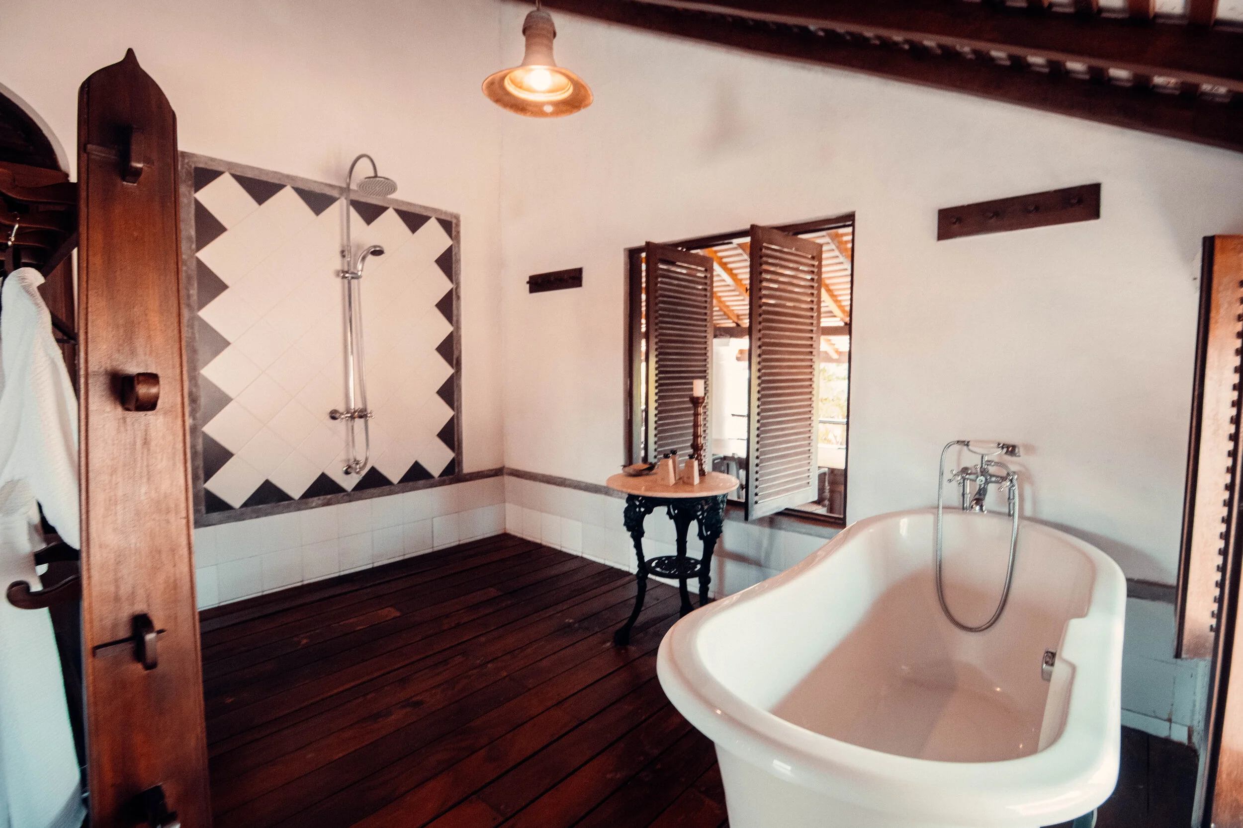 A rustic bathroom with dark wooden flooring, a vintage clawfoot bathtub, a wall-mounted shower, and open window shutters allowing natural light.
