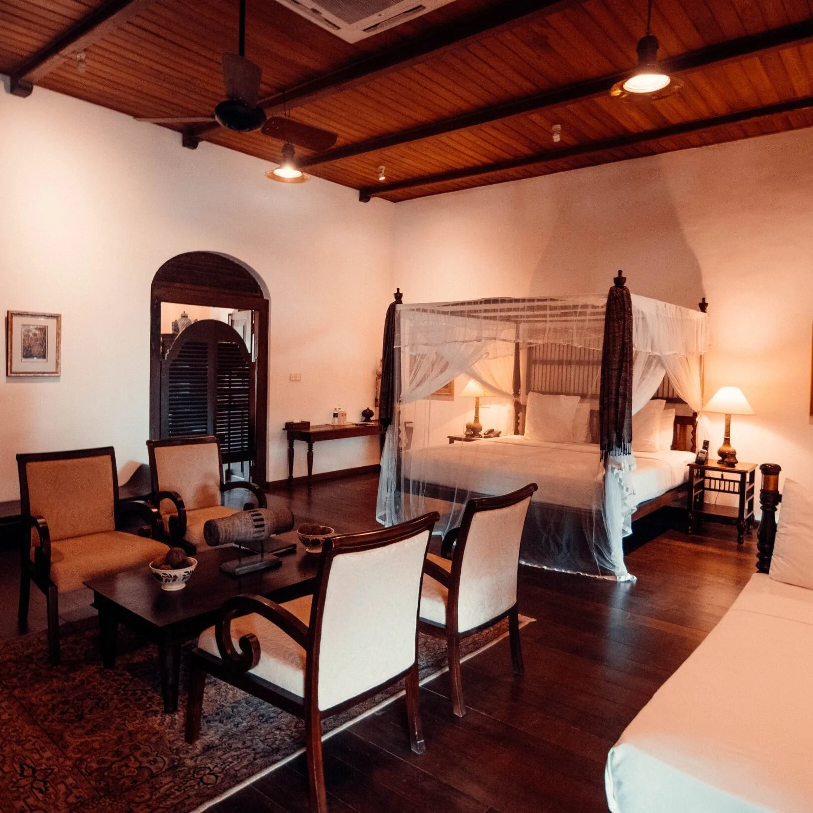 A hotel room with a dark wood four-poster bed and canopy, a seating area with chairs and a table, and warm lighting in a rustic setting.