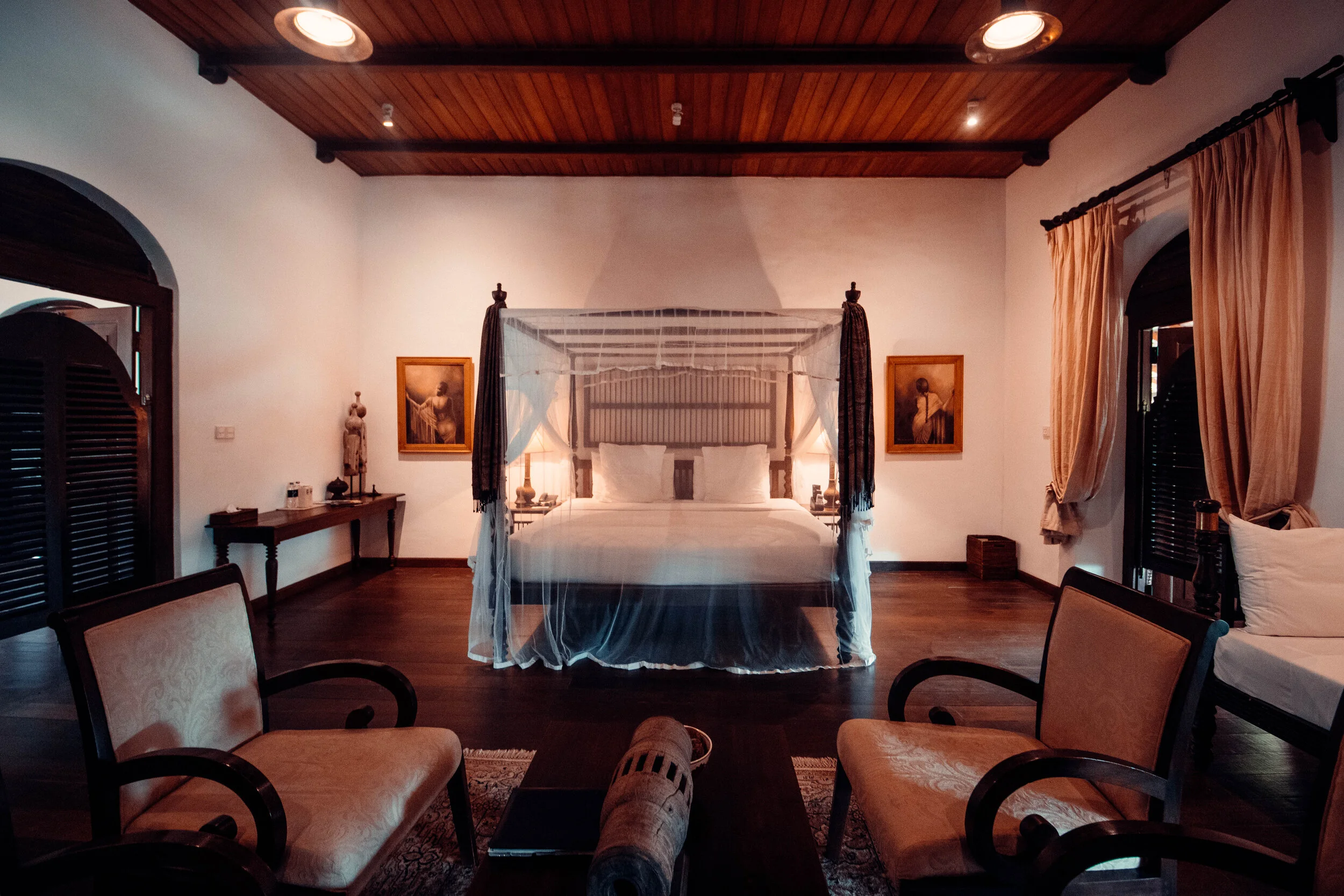 A bedroom with a four-poster bed in the center, draped with a mosquito net. Two framed pictures hang on the wall behind the bed. There are two nightstands with lamps on either side of the bed. Two armchairs are positioned in front of a wooden coffee 