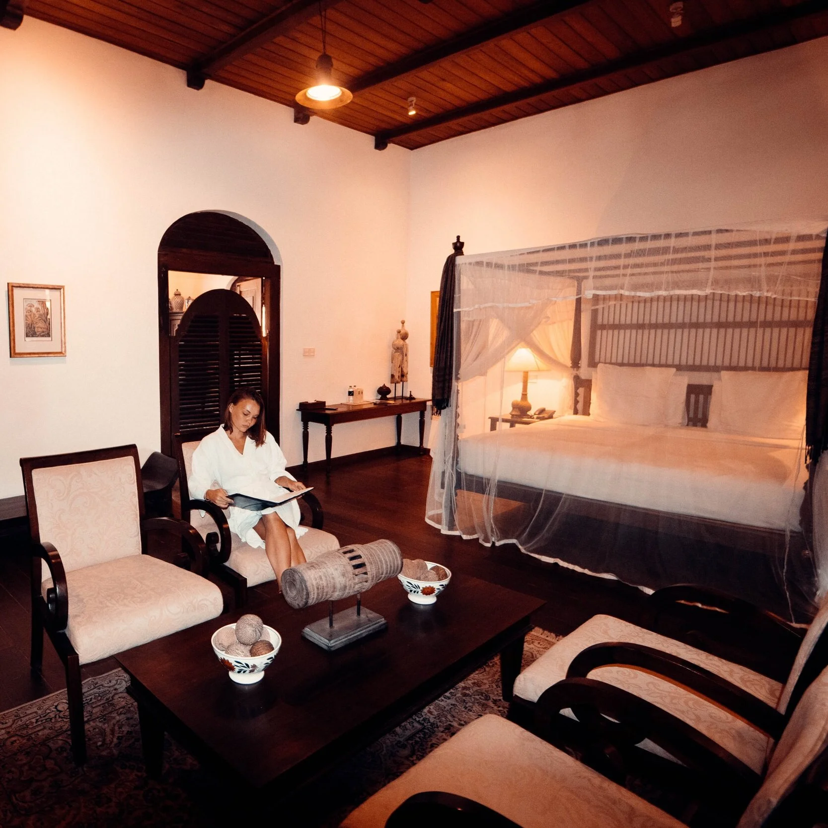 A woman dressed in a white robe sitting on a beige armchair in a hotel room, reading a book. The room has dark wooden furniture, a large four-poster bed with a mosquito net, a nightstand with a lamp, and decorative items on a long table against the w