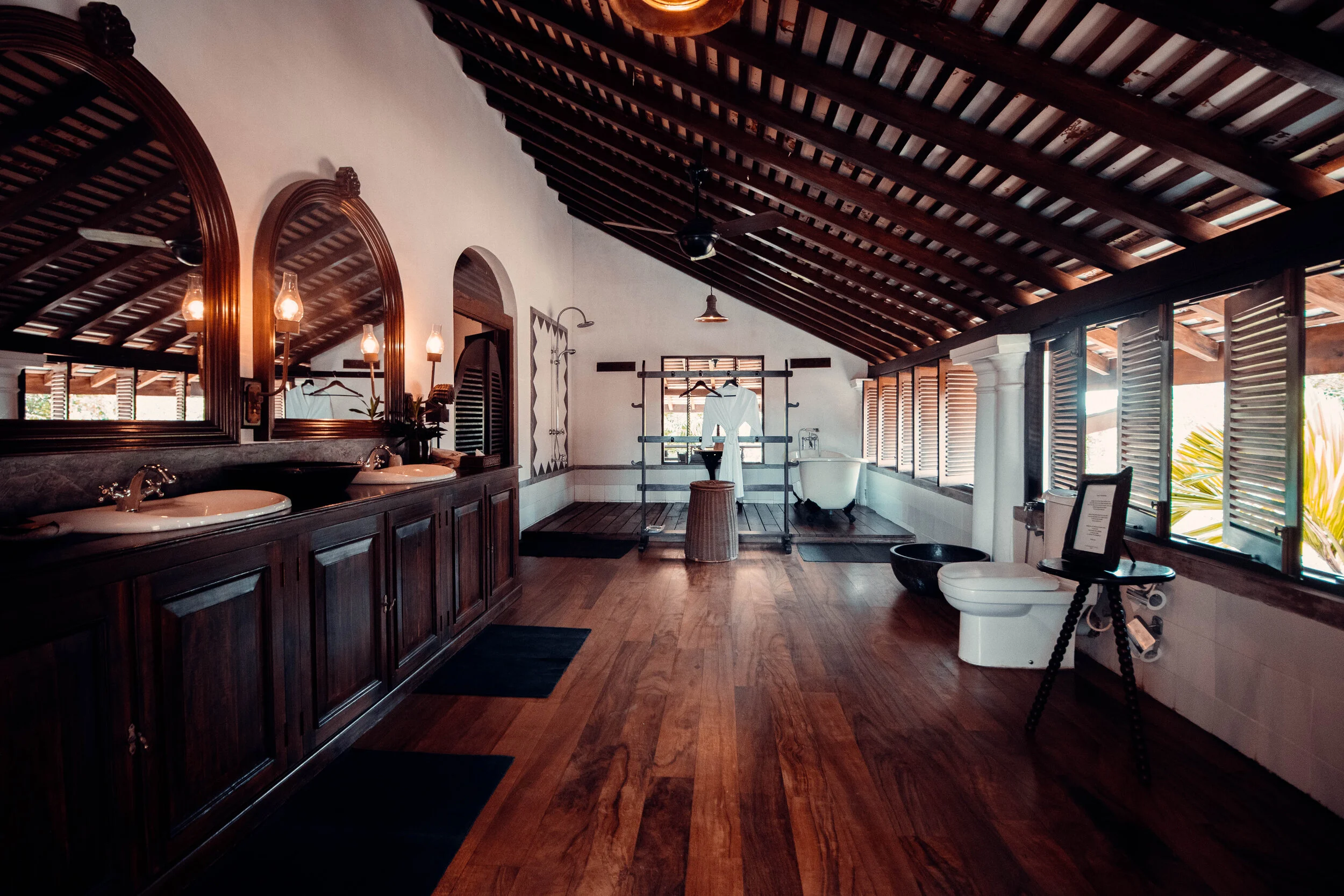 Spacious bathroom with wooden floors and sloped wooden ceiling, featuring two sinks with mirrors, a vintage bathtub, a toilet, large windows with wooden shutters, and wooden furniture and decor.