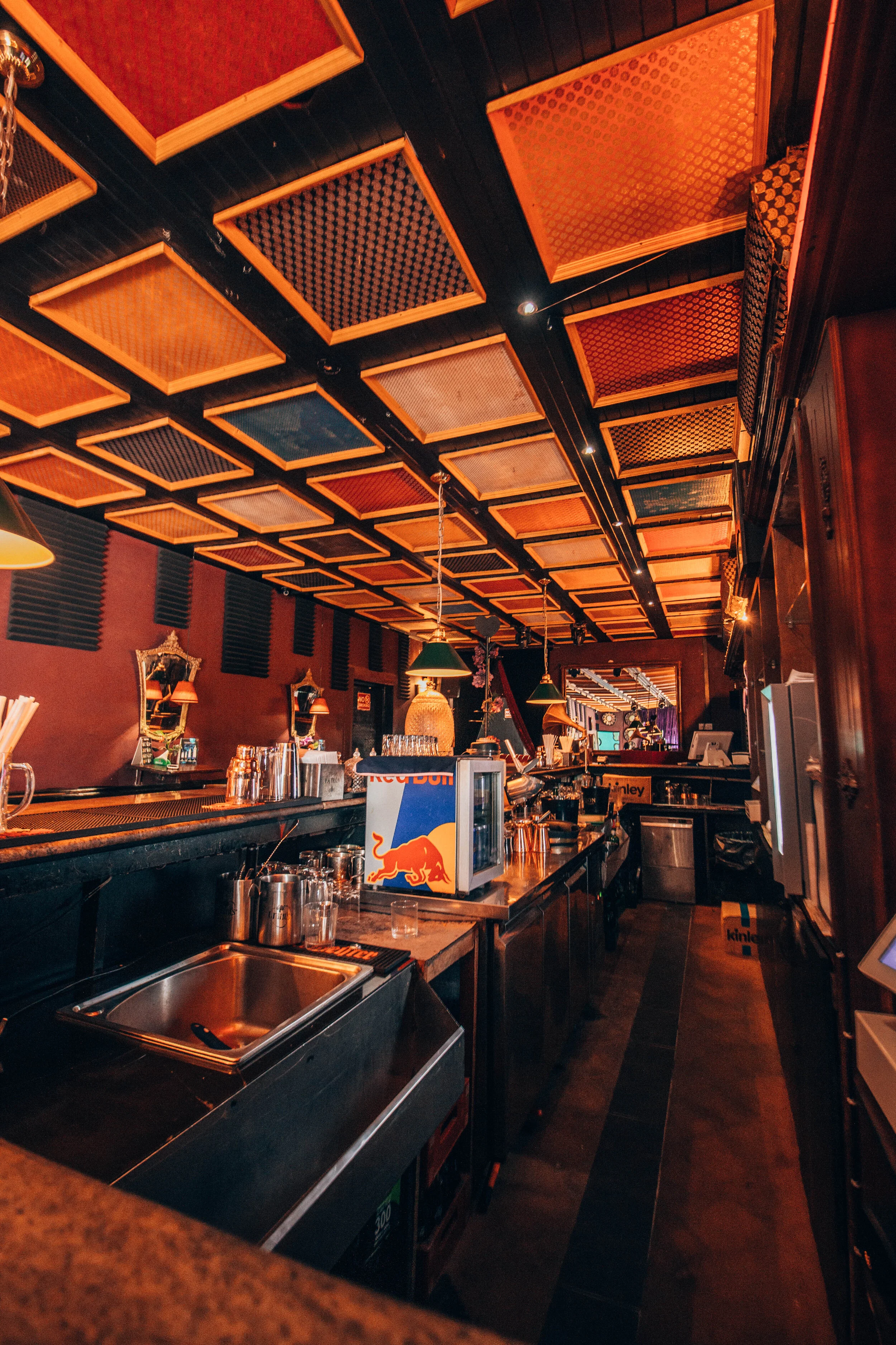 Interior of a bar or restaurant with a wooden and orange color theme, featuring patterned ceiling panels, green pendant lights, and various bar equipment.