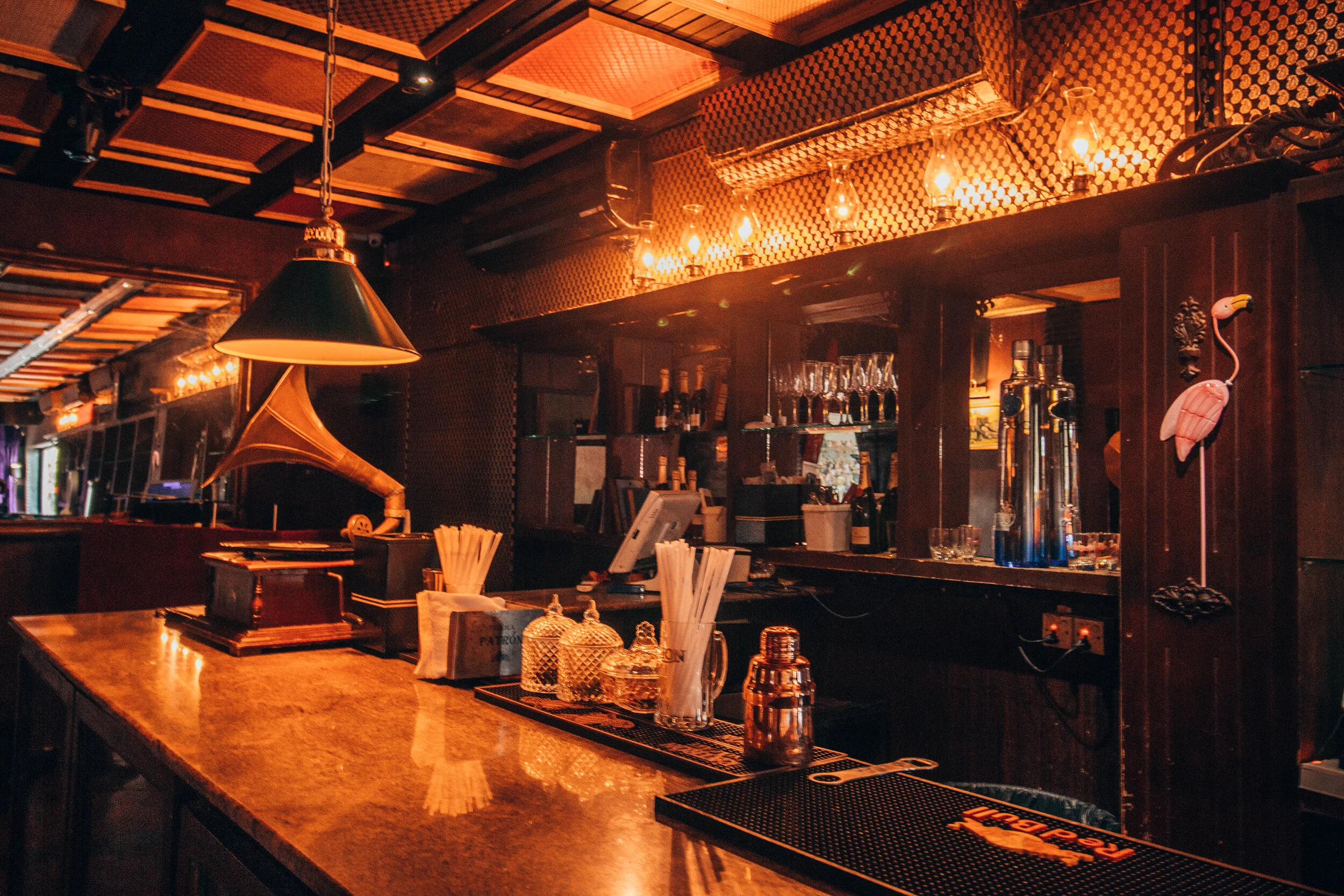 Interior of a dimly lit bar with a wooden counter, glasses, bottles, and vintage decor including a flamingo lamp and a gramophone.